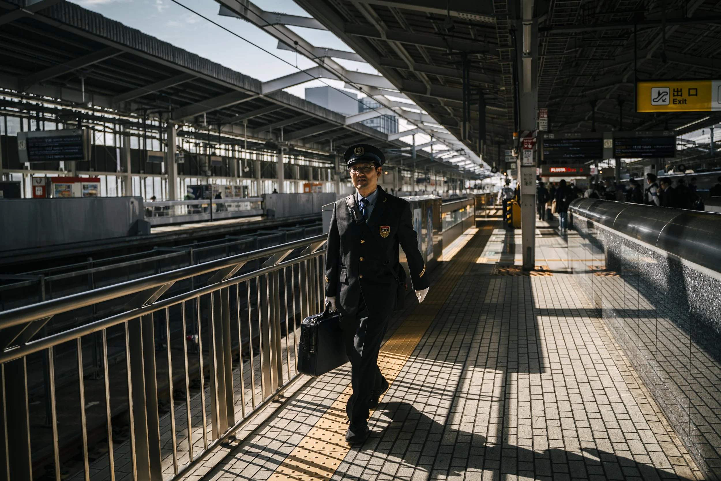A man in a uniform and cap walking at an outdoor train station, pulling a suitcase, with other travelers in the background and signs indicating an exit and platform information.