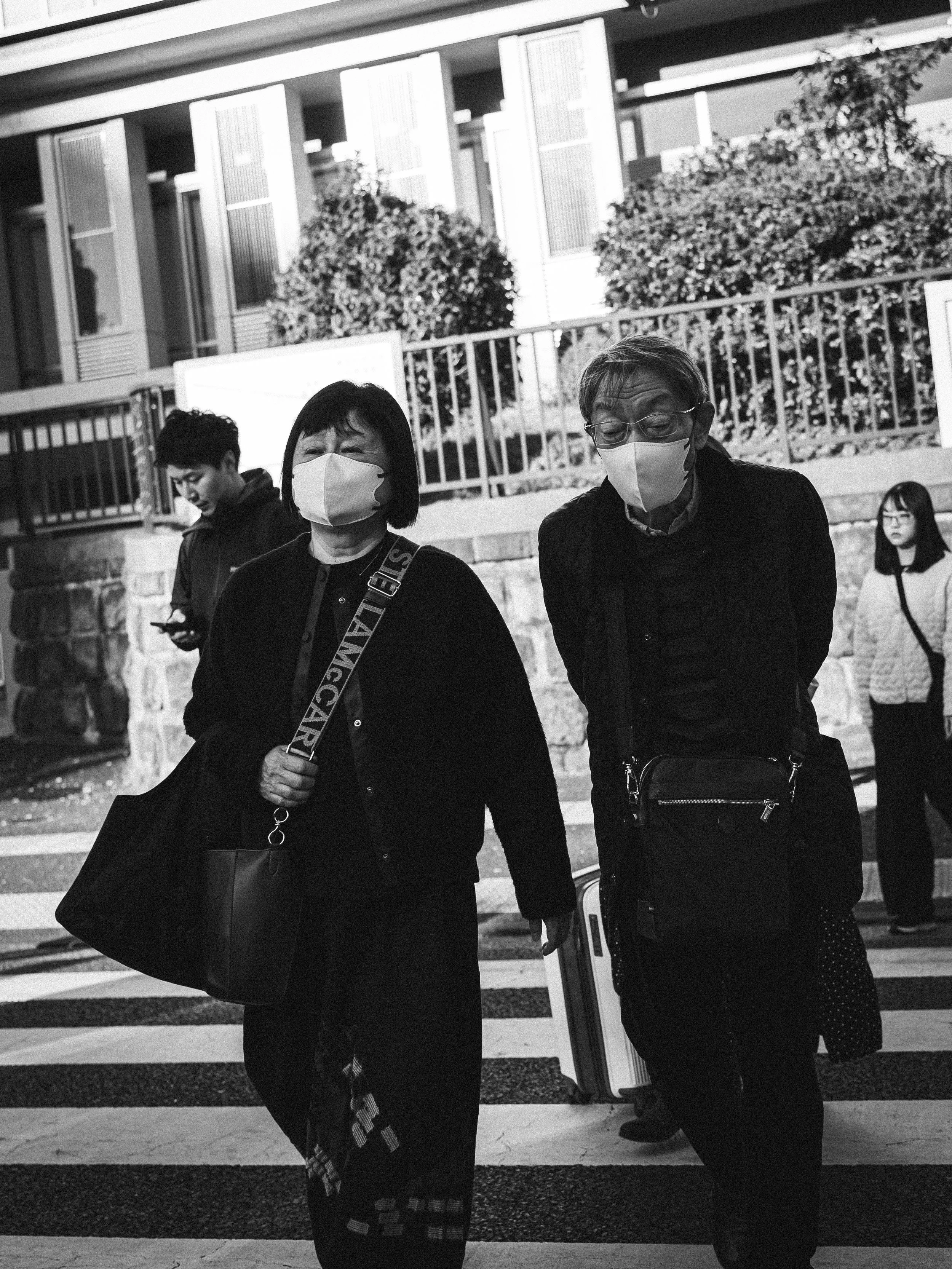 Black and white photo of three adults and one child at a crosswalk in an urban area. The two adults in the foreground are women wearing masks, one carrying a purse and the other holding a bag, and the child in the background is a girl wearing glasses