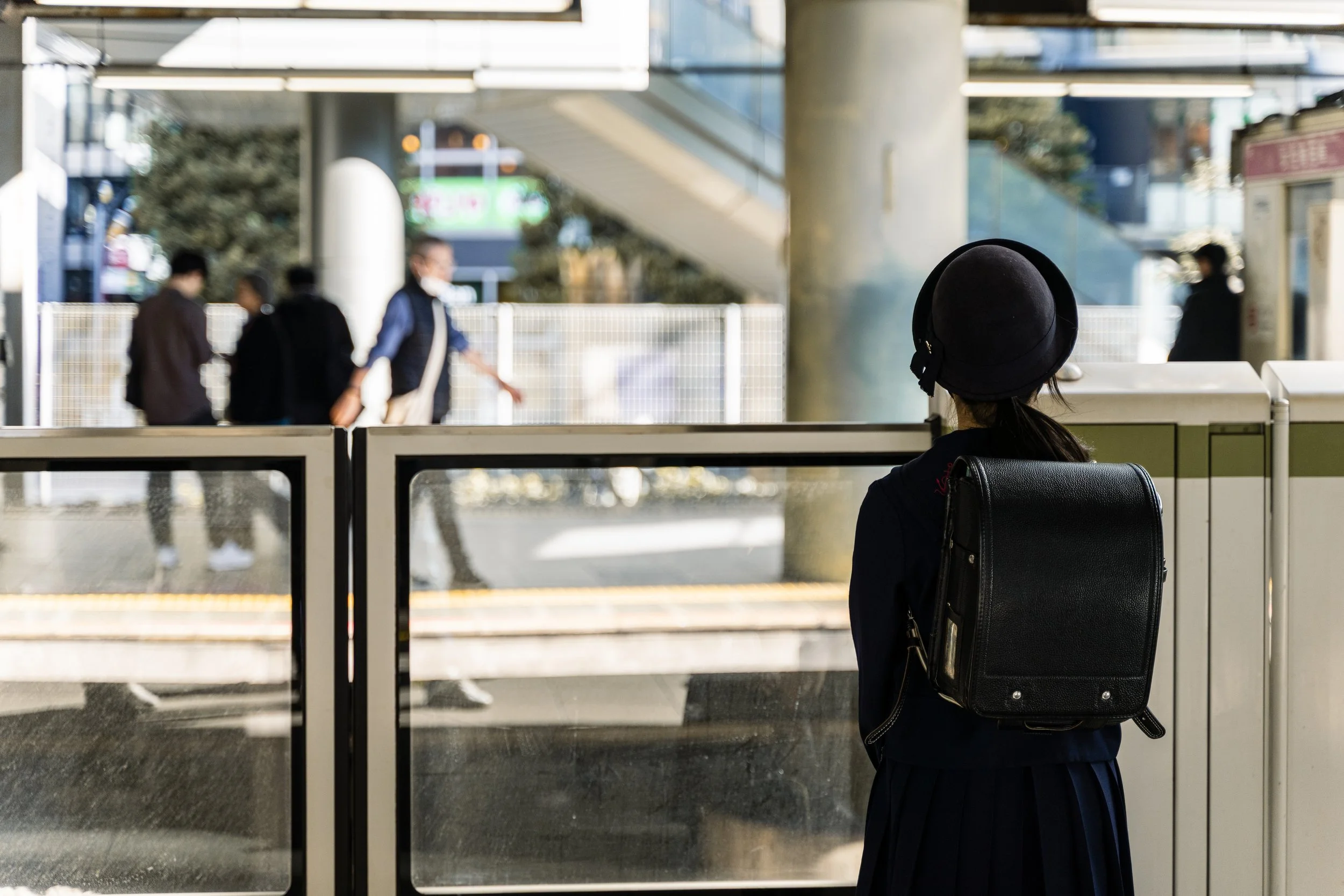 A woman wearing a black hat and carrying a black backpack, waiting at a train station or airport terminal, looking out through glass doors. In the background, people are walking outside, some wearing face masks.