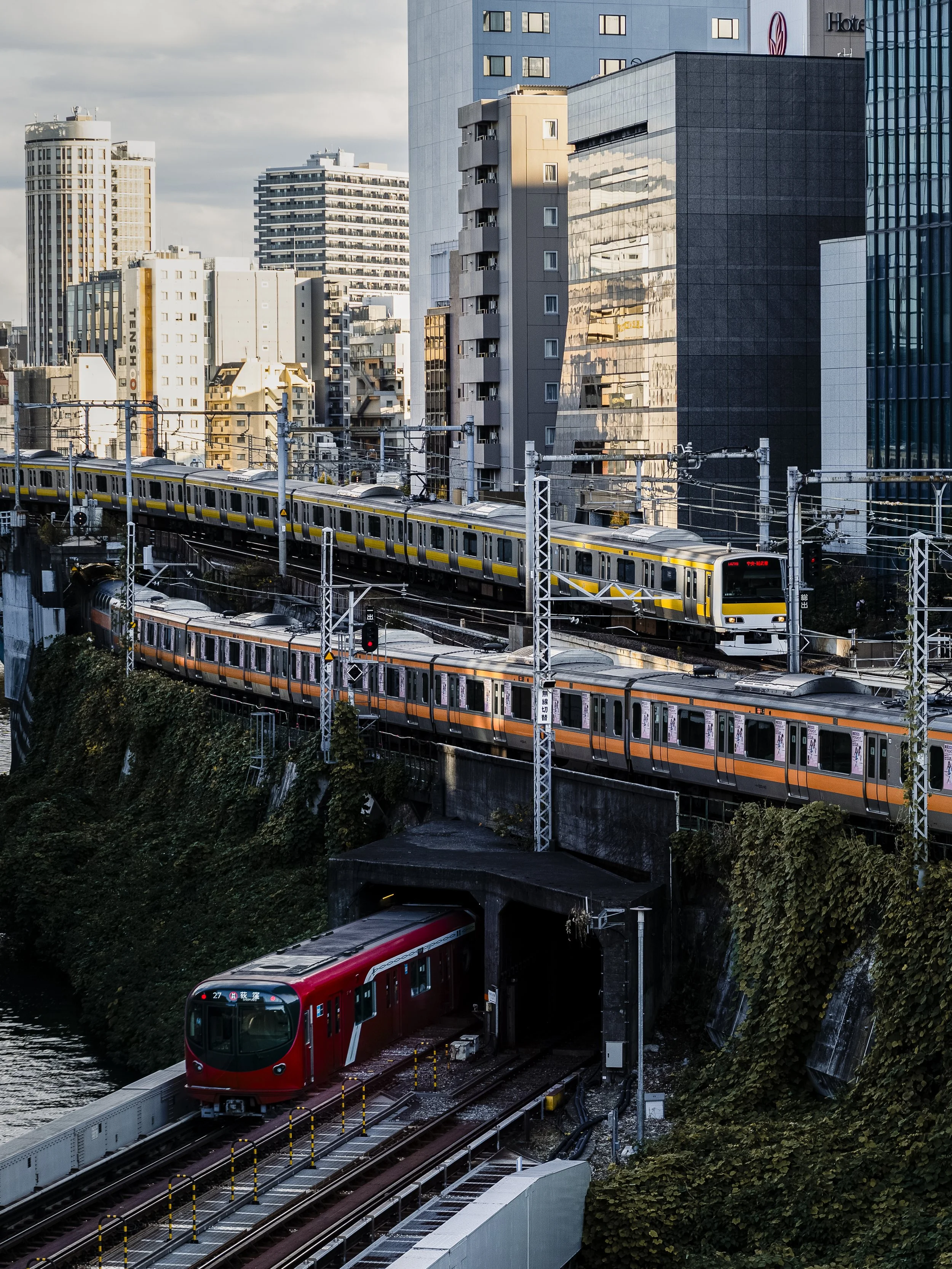 Ochanomizu, Tokyo — Three trains, three lines, three colours — yellow Sobu, orange Chuo, red Marunouchi — caught simultaneously above the Kanda River. Tokyo's rail network at its most layered. The city as a machine, and the trains its moving parts.

