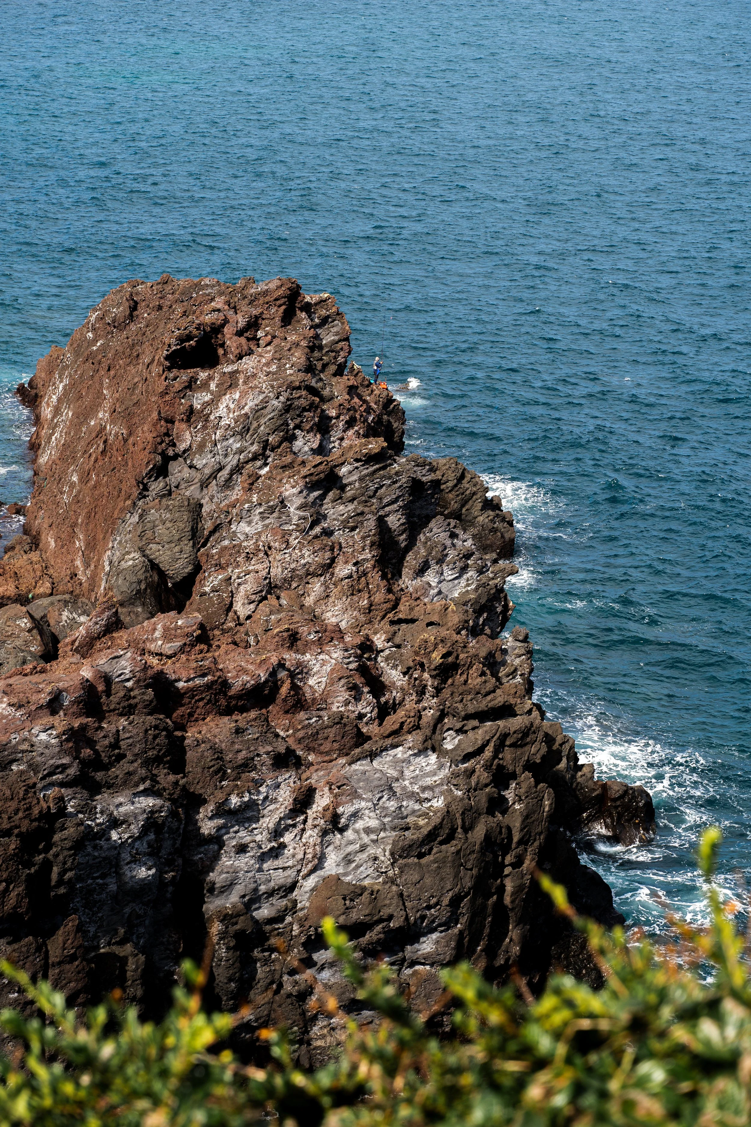 Jeju Island, South Korea — Two figures at the edge of a volcanic cliff, lines cast into the sea below. The sea doesn't notice them. They don't seem to mind.