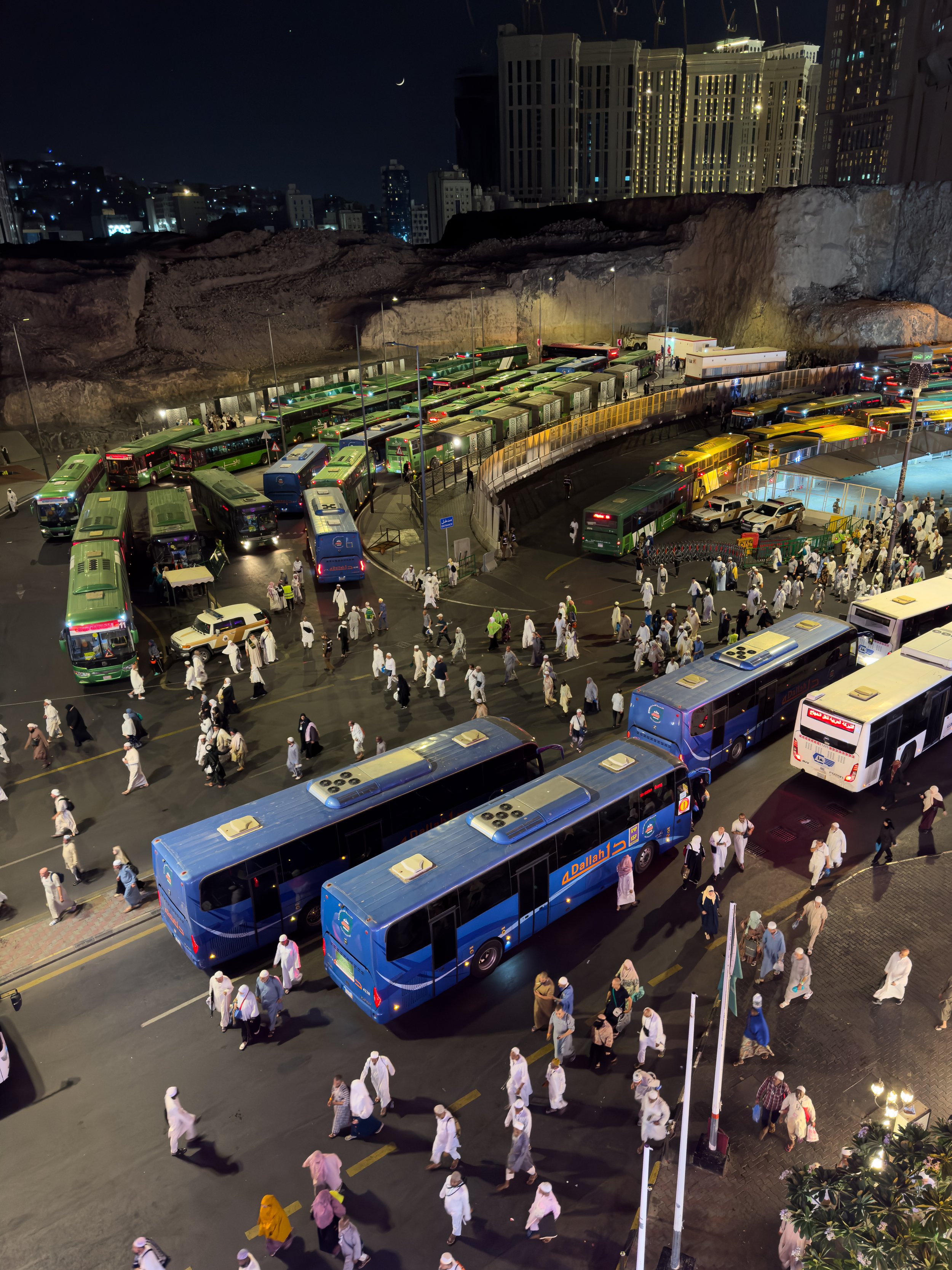 Mecca, Saudi Arabia — A bird's eye view of the pilgrim transport hub at night — buses, crowds and the crescent moon above. The logistics of moving millions, seen from above. Organised chaos in service of faith.