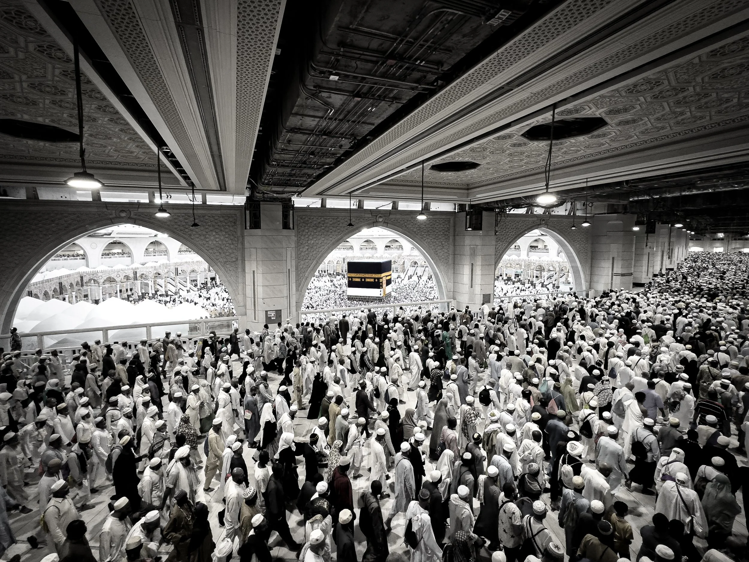 A crowded Islamic pilgrimage inside a large ornate mosque with many men wearing white clothing and hats, gathered around the Kaaba, visible through an archway.