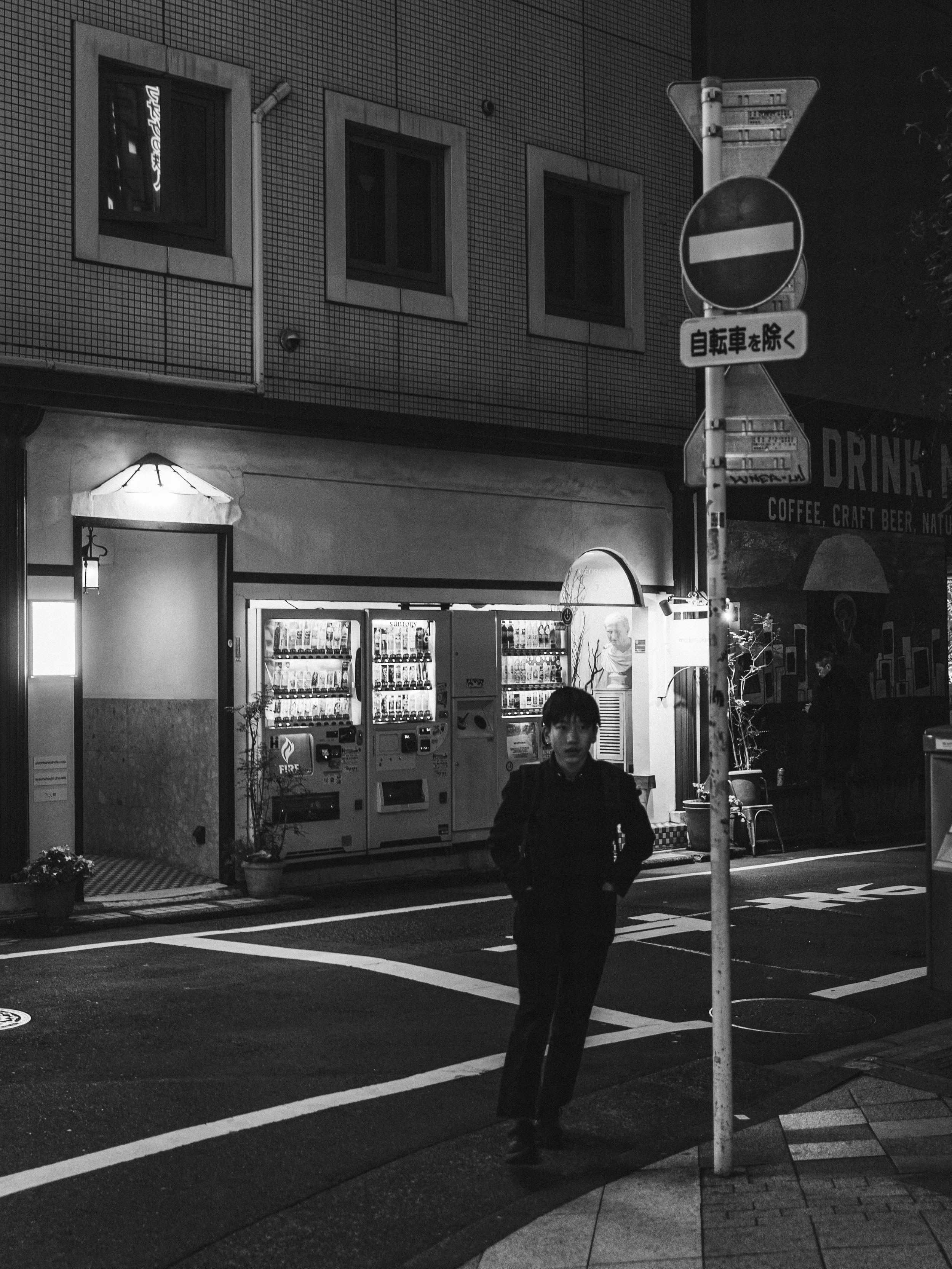 A person walking on a city street at night near a vending machine and street signs. The scene is in black and white.
