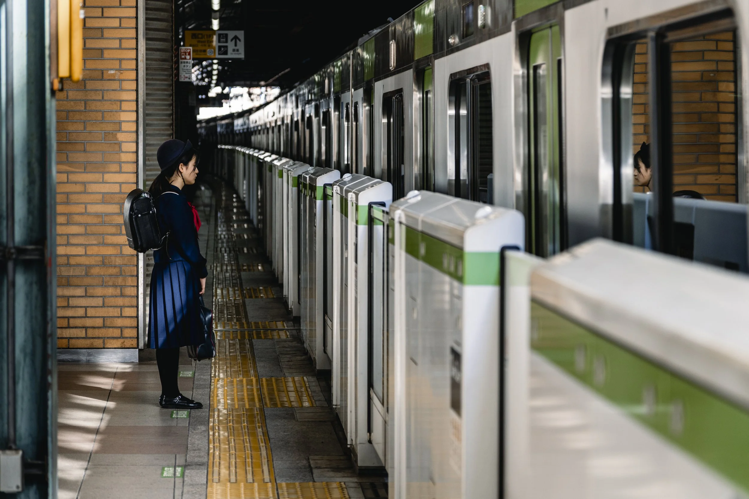 A young woman in a blue uniform and hat, carrying a backpack, waits near train ticket gates at an indoor train station.