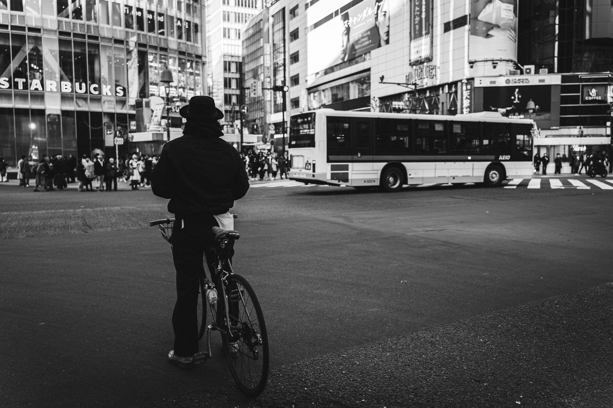 A person with a bicycle standing on a city street corner in front of a crosswalk, with a busy urban environment and tall buildings in the background.