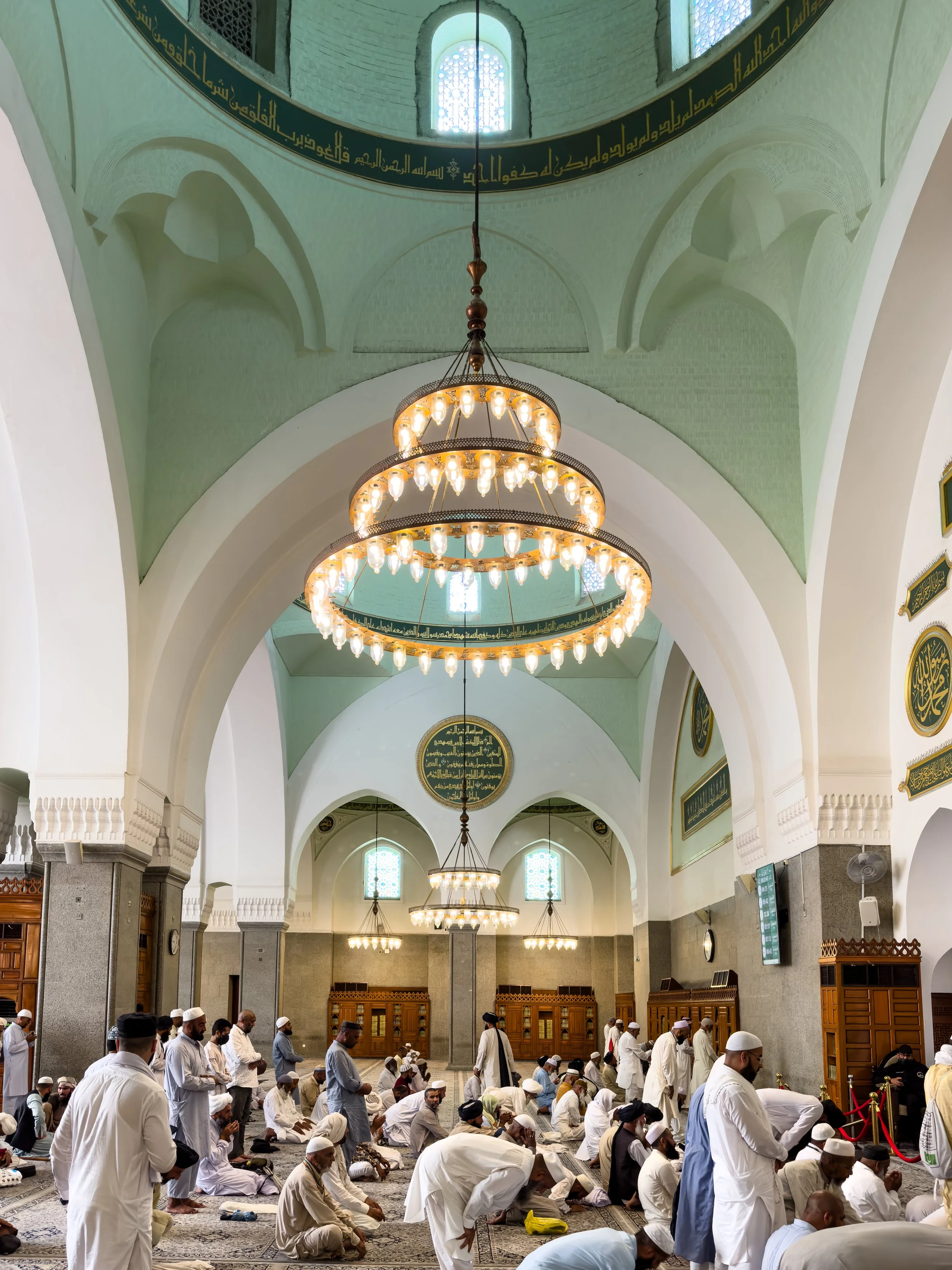 Masjid Quba', Medina, Saudi Arabia — Pilgrims gather beneath an ornate chandelier in one of the Prophet's Mosque's oldest halls. The arched ceiling, the warm light, the white robes — a stillness that exists even in the middle of millions.