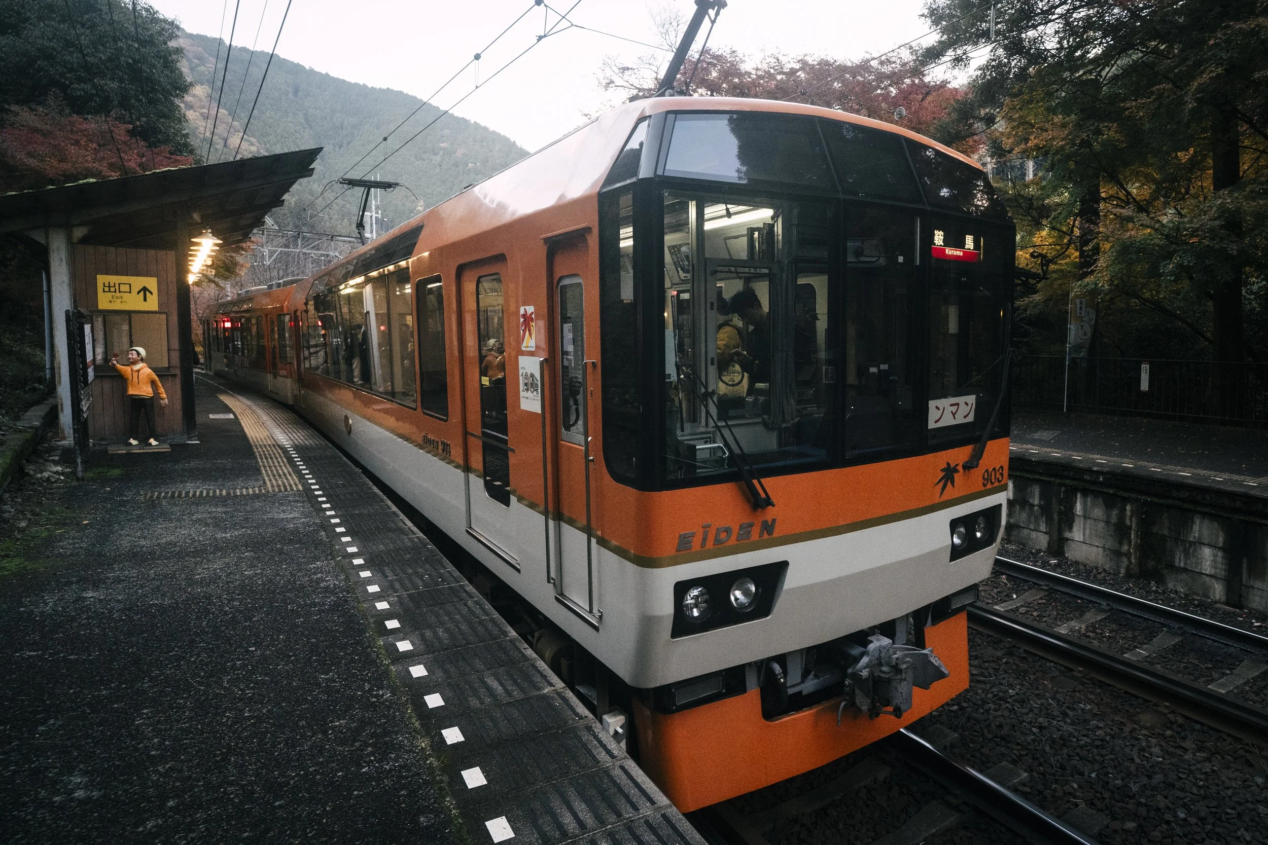 Eizan Dentetsu 903, Kurama, Kyoto — The orange Eiden pulls into Kurama station as autumn closes in around the platform. A lone figure in a yellow jacket at the exit. The end of the line made beautiful by the season.