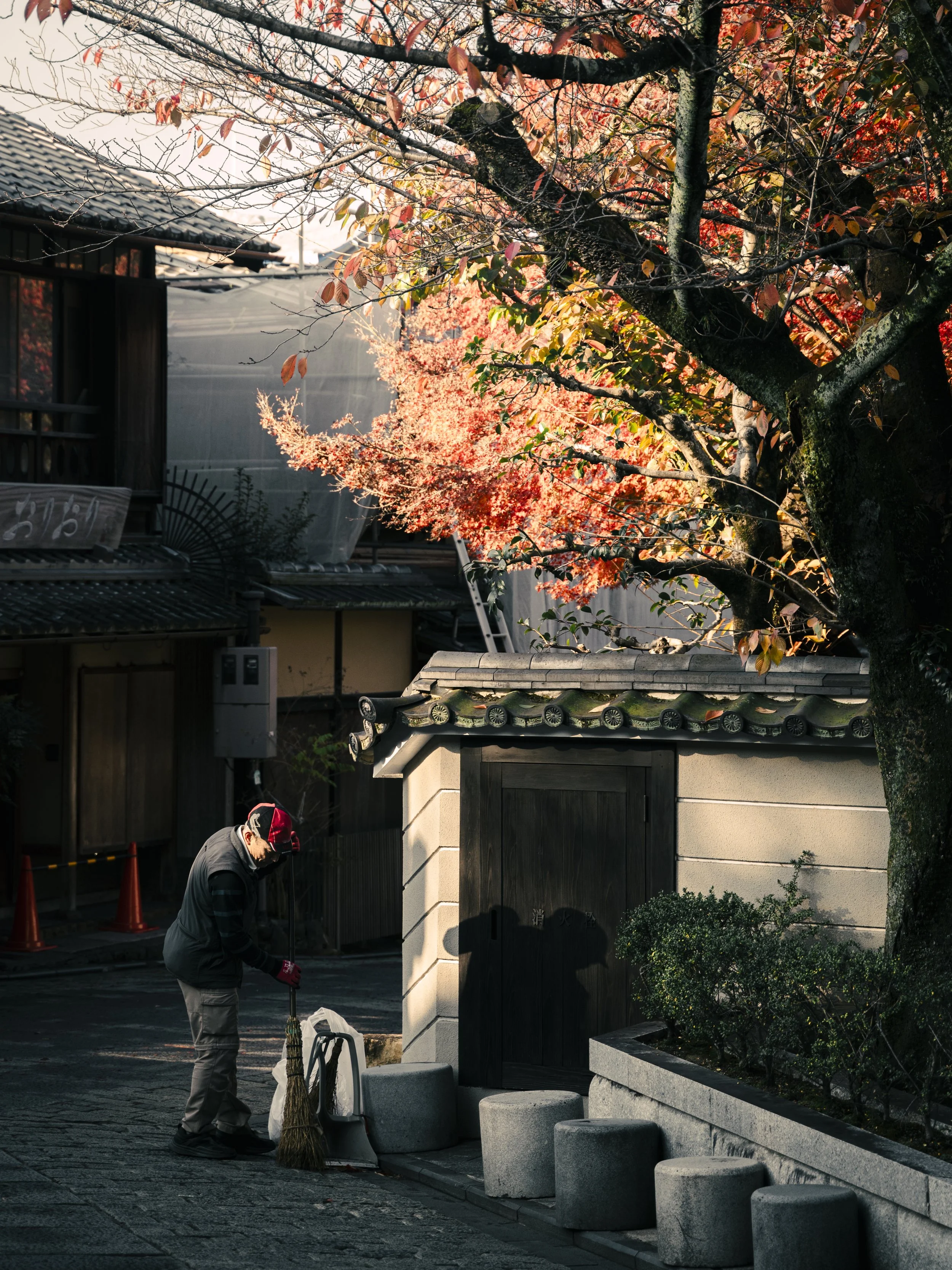 A man cleaning the sidewalk in front of a building with traditional Japanese architecture, including a tiled roof and wooden gates, in autumn. There are orange traffic cones and orange leaves on the tree.