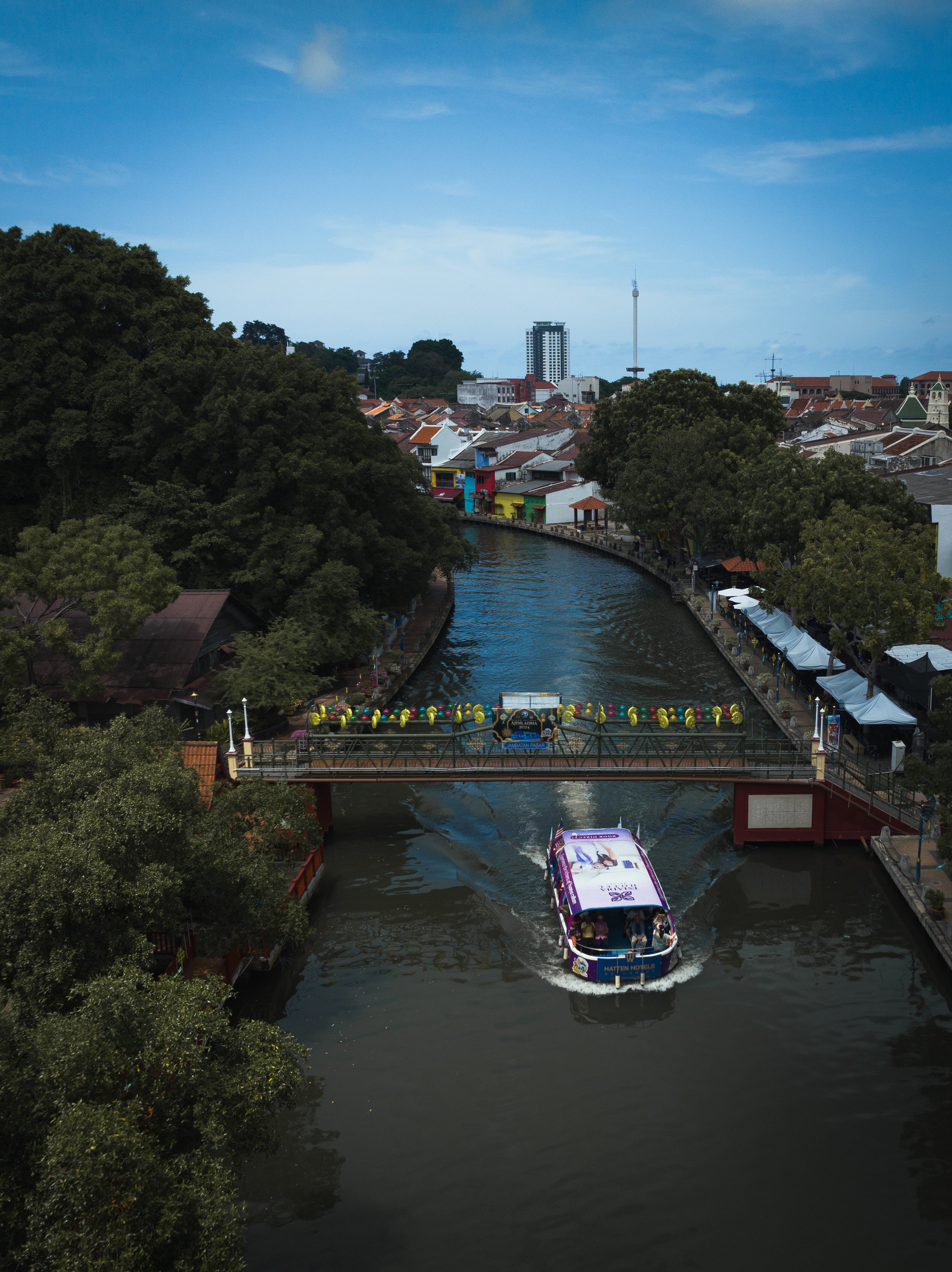 Sungai Melaka, Melaka, Malaysia — A river cruise passes under a decorated pedestrian bridge as the old city unfolds along both banks. The Melaka Tower watches from the distance. Four centuries of history, still very much in use.
