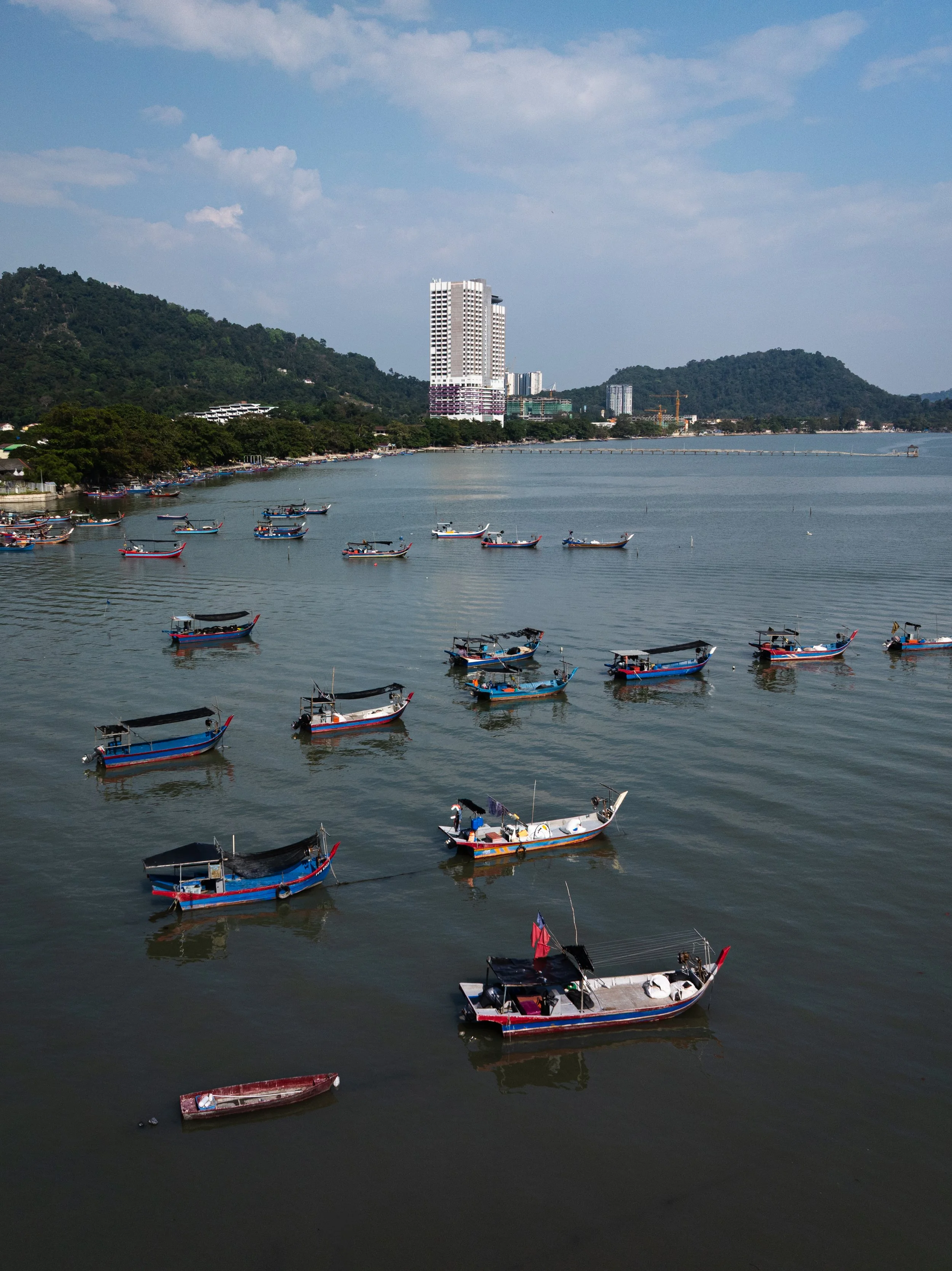 Penang, Malaysia — Fishing boats scattered across the bay in the afternoon light, the forested hills of Penang rising behind. Working vessels on working water — the other side of an island most people only see from the beach.