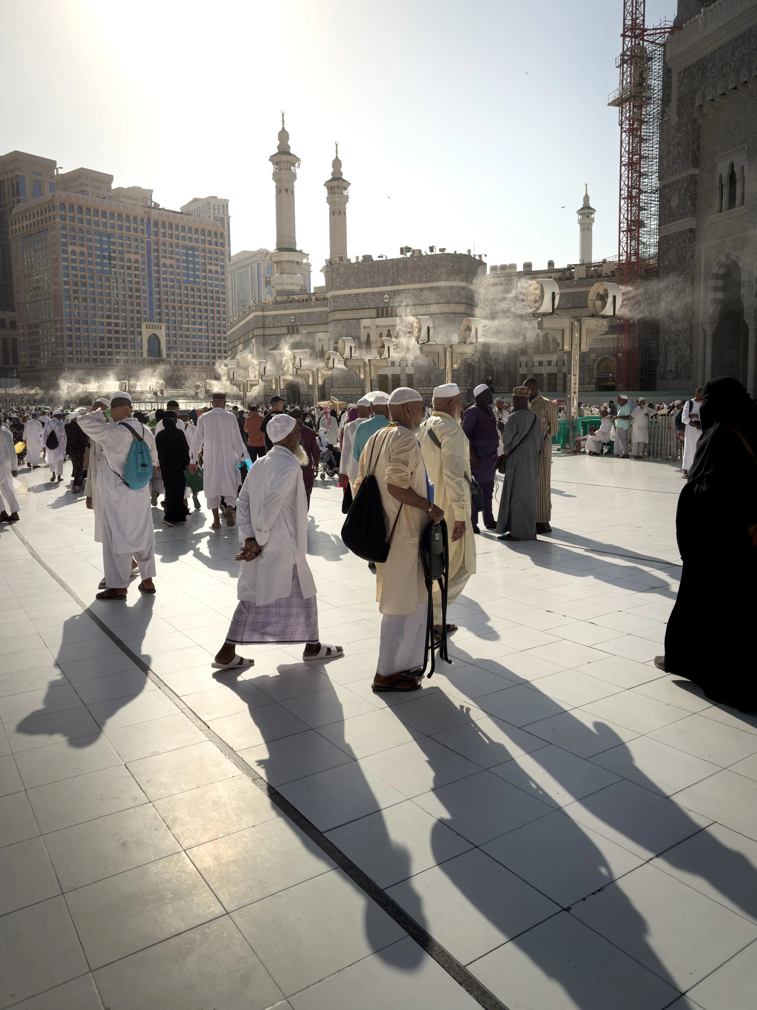 Masjidil Haram, Mecca, Saudi Arabia — Long shadows stretch across the white marble courtyard as pilgrims move through the late afternoon heat, mist fans softening the air behind them. Light and shadow and the quiet determination of those still walkin