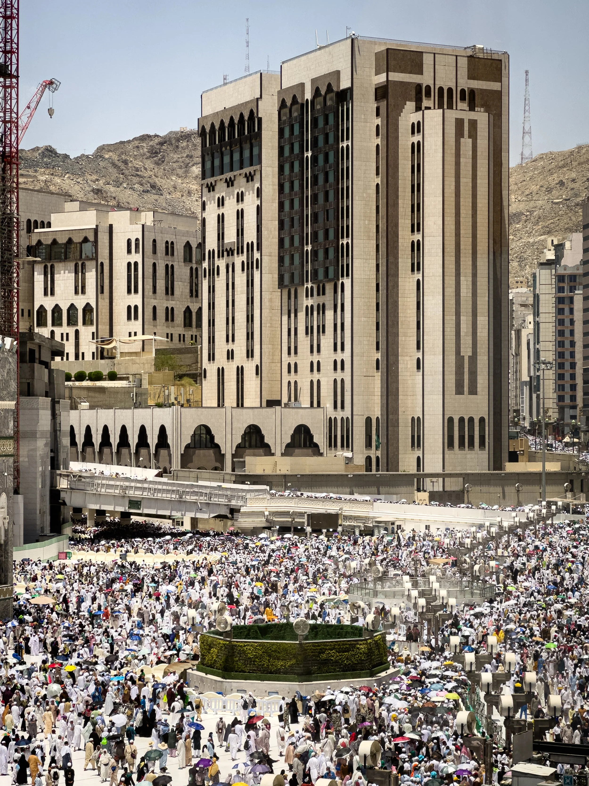 Masjidil Haram, Mecca, Saudi Arabia — The outer courtyard fills with thousands of pilgrims between prayers, the Ajyad hills rising behind the surrounding buildings. From above, individual faces disappear into a sea of white. The scale of Hajj, in a s
