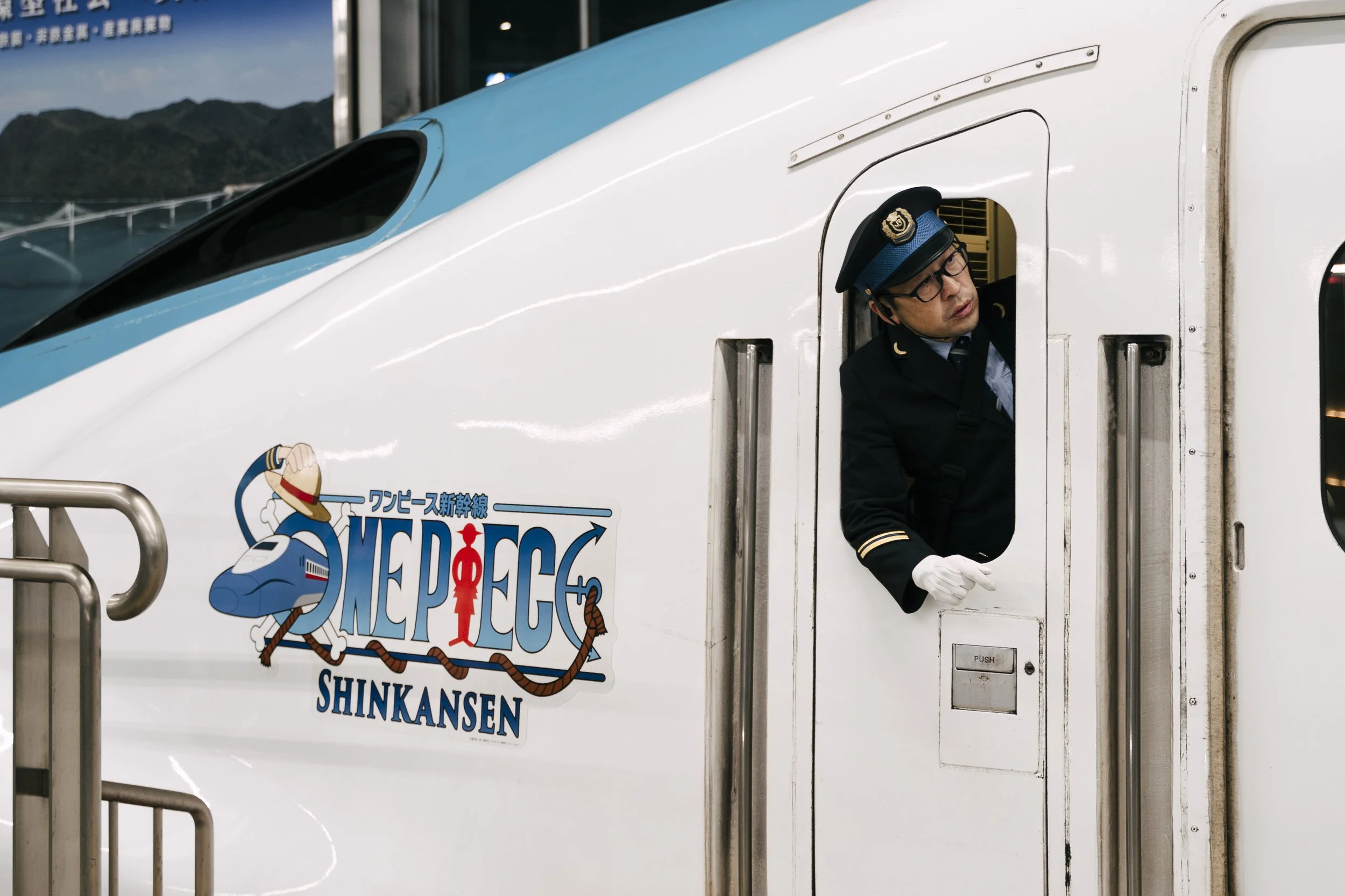 A train conductor wearing a dark uniform, glasses, and a cap leaning out of a train window. The train has a colorful sign with a cartoon character and the words "ONE PIECE SHINKANSEN" on the side, with mountains visible in the background.