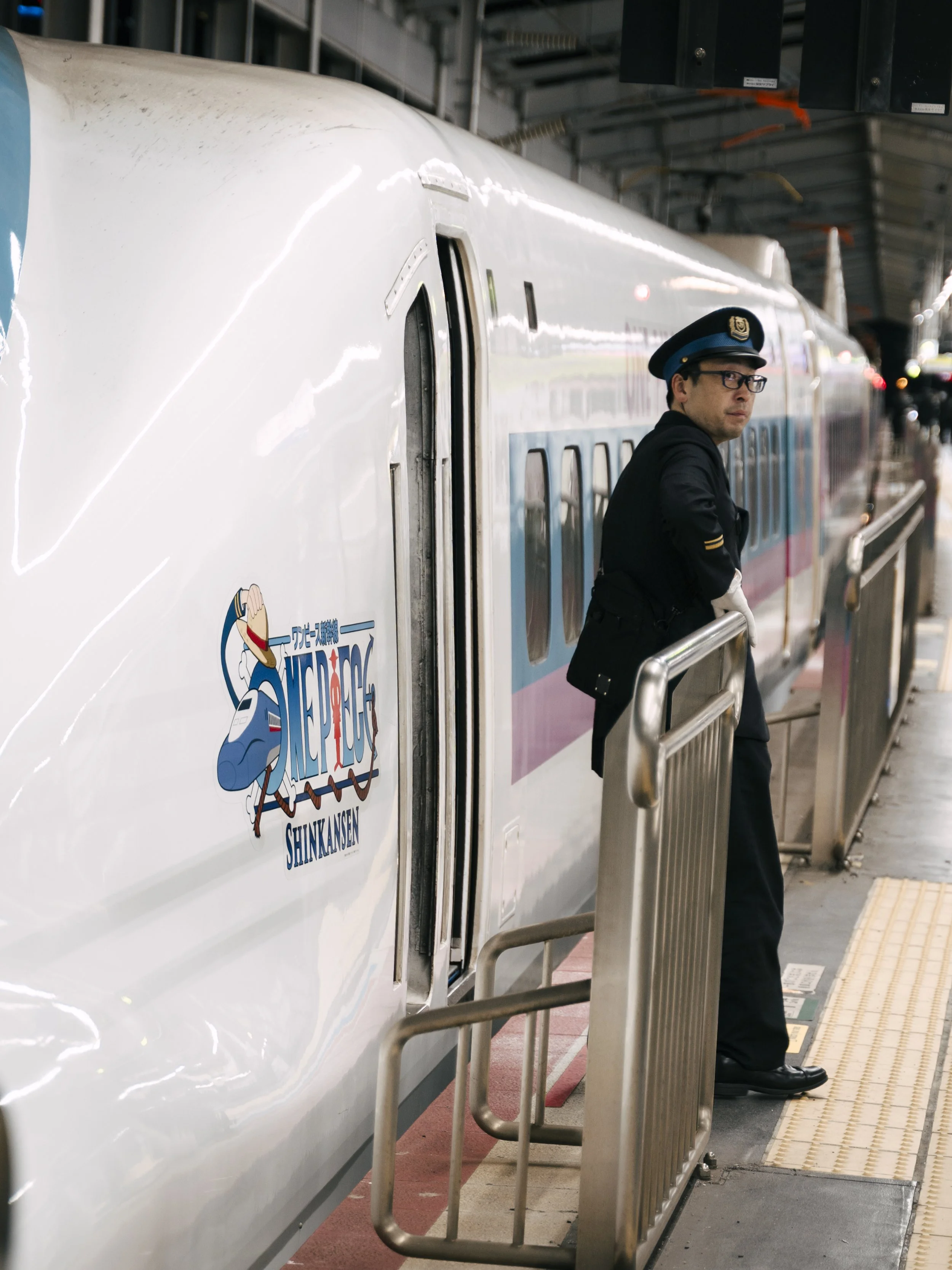 A person dressed as a train conductor or railway staff standing on a train platform beside a Shinkansen bullet train, wearing a navy uniform and cap, looking towards the camera.