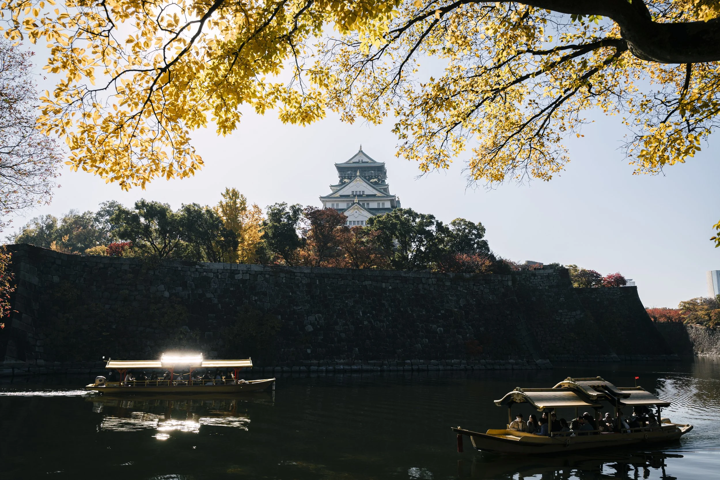 Osaka Castle, Japan — Wooden boats drift across the moat as autumn turns the trees gold above four centuries of stone. The boats still run.