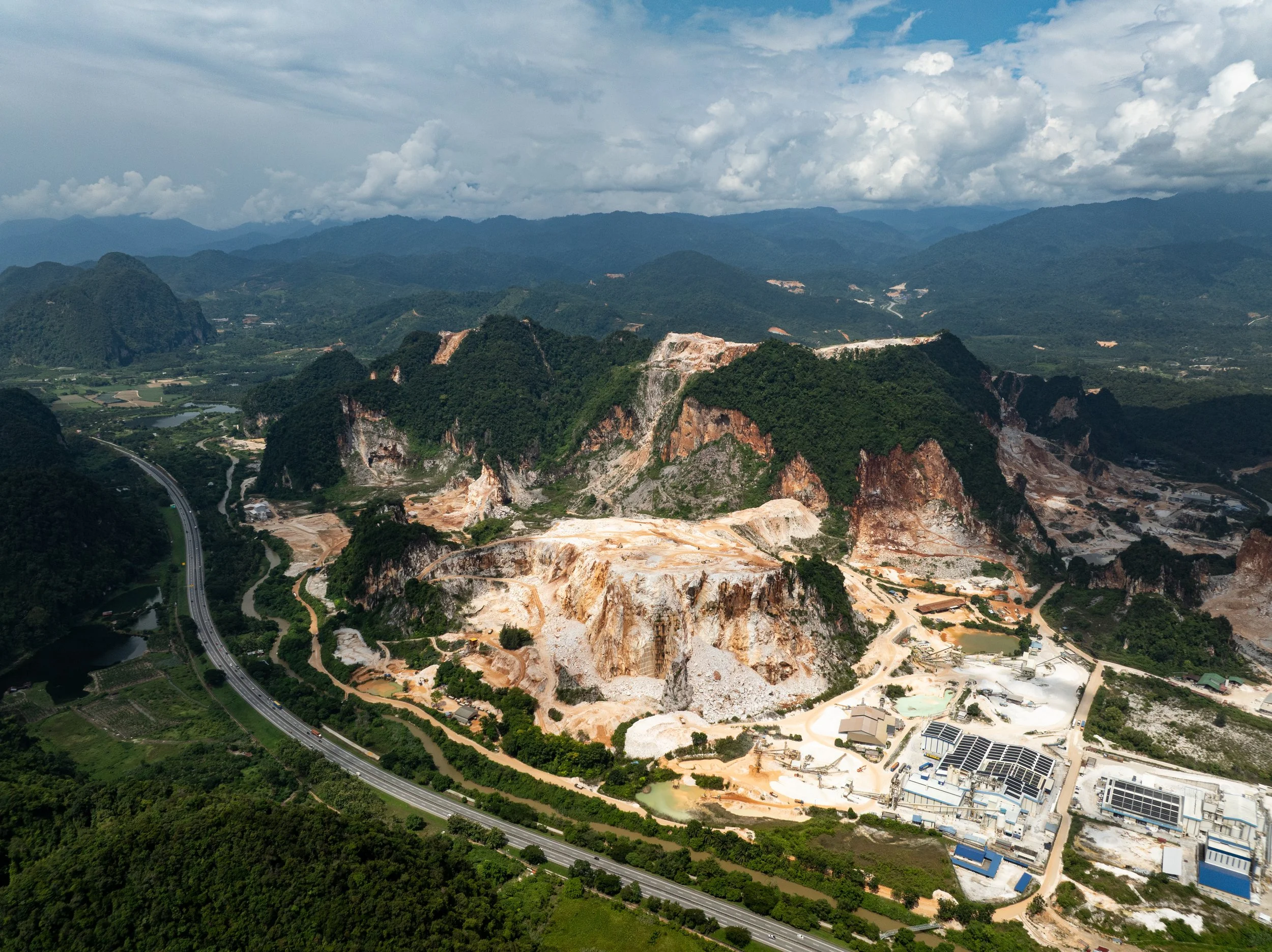 Wide aerial perspective of large-scale limestone quarrying operations alongside a major highway in Simpang Pulai, Ipoh, Perak.