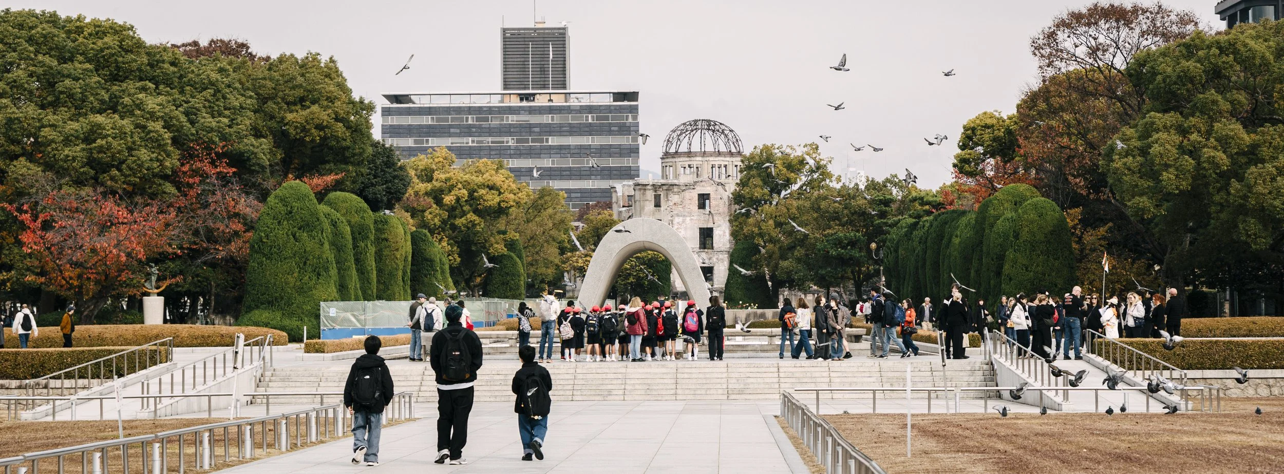 Tourists visiting the Hiroshima Peace Memorial Park, with the Atomic Bomb Dome in the background, surrounded by autumn trees and pigeons flying.