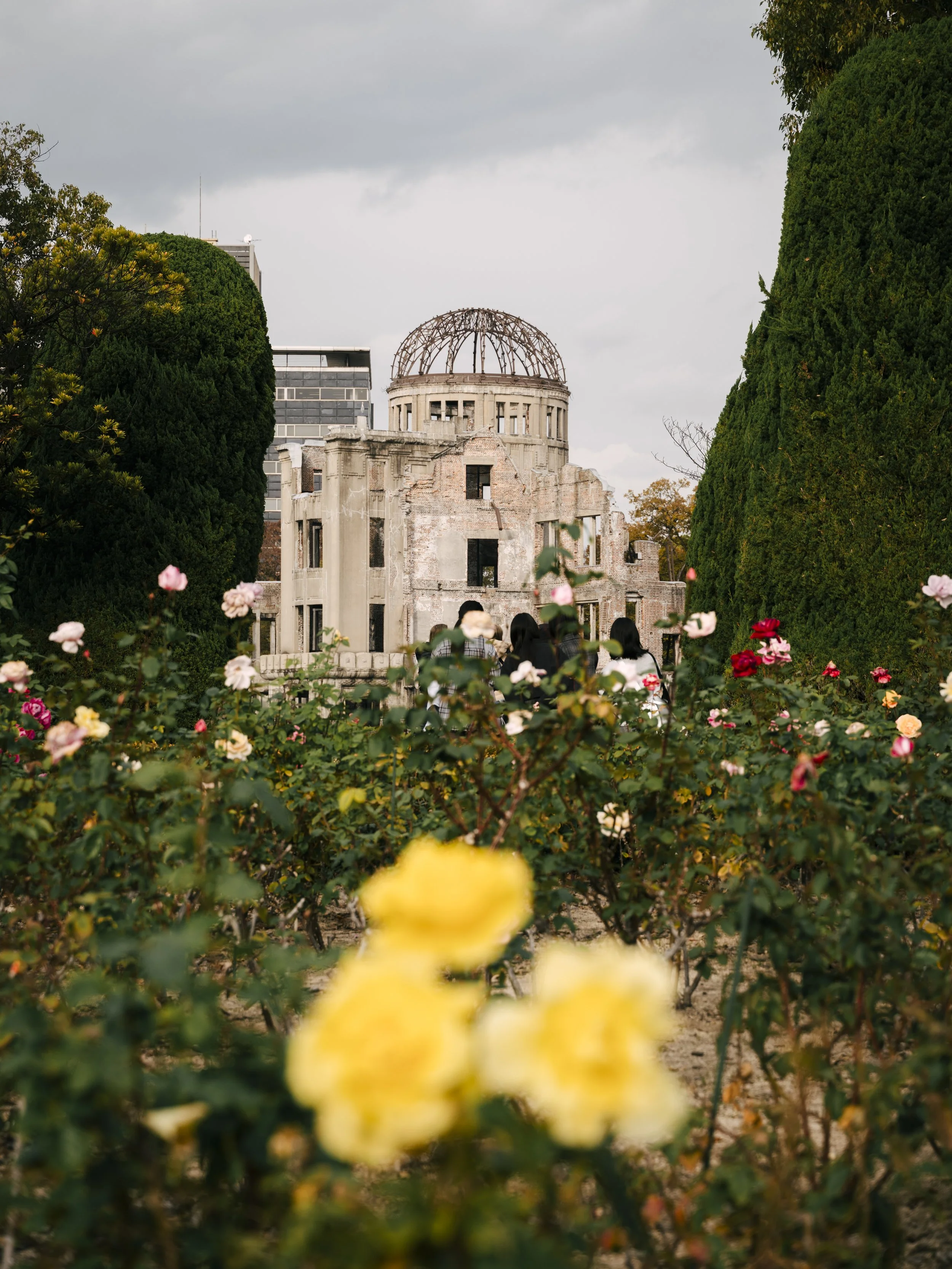 Ruins of the Hiroshima Peace Memorial with flowering bushes in the foreground, and a cloudy sky in the background.
