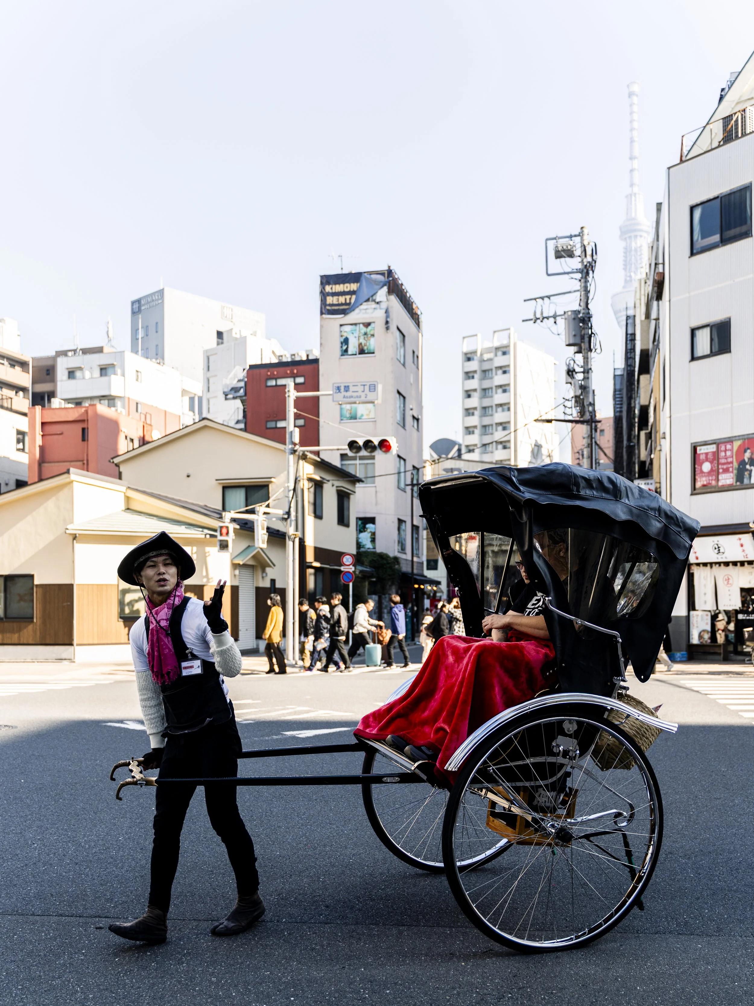 A street scene with people and buildings in the background. A person wearing a black hat, beige sweater, and pink scarf is walking near a rickshaw with a red blanket in it. The rickshaw is on the street next to the pedestrian.