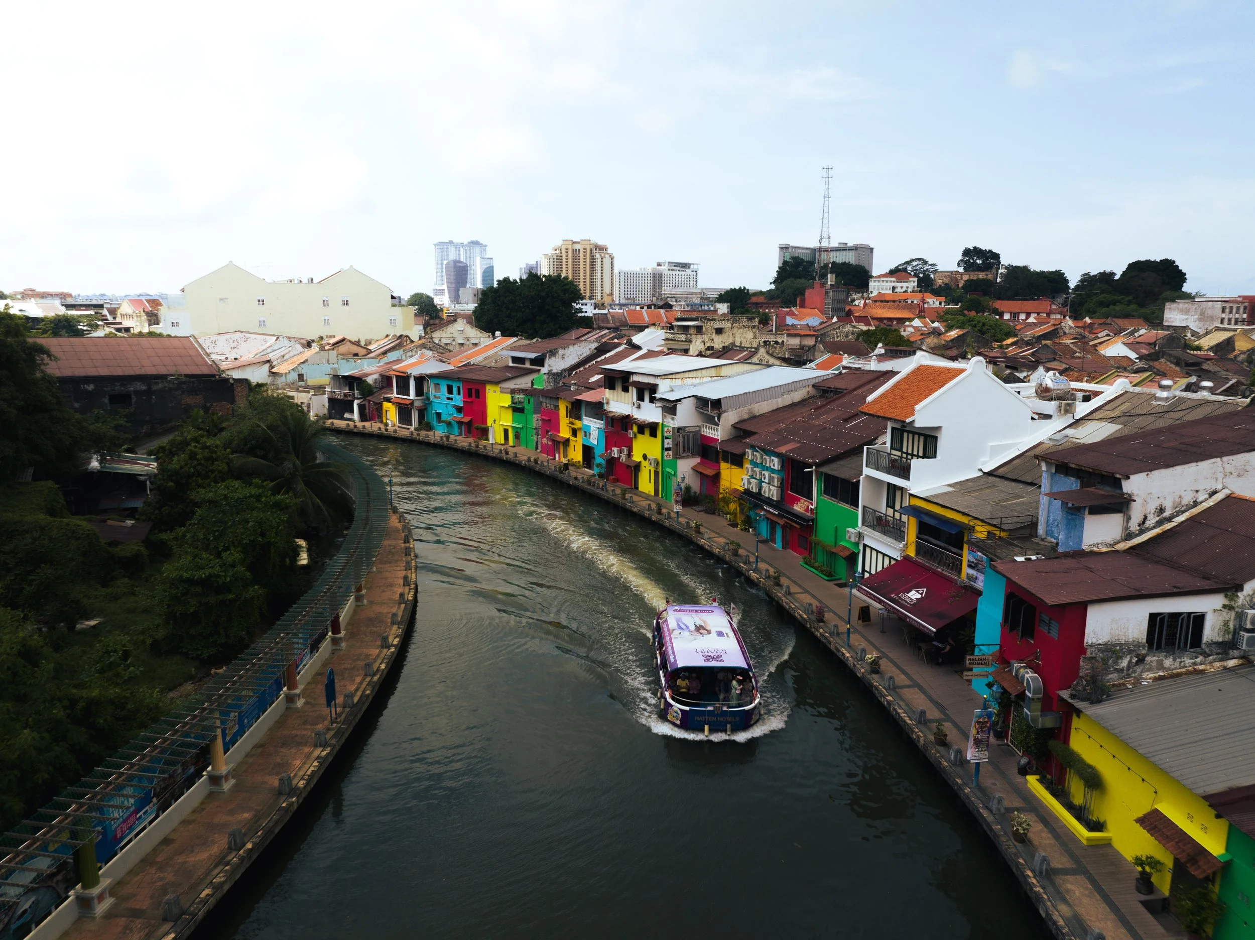 Sungai Melaka, Melaka, Malaysia — A river cruise winds past a row of colourful heritage shophouses, the old city stacking up behind in layers of terracotta and history. 