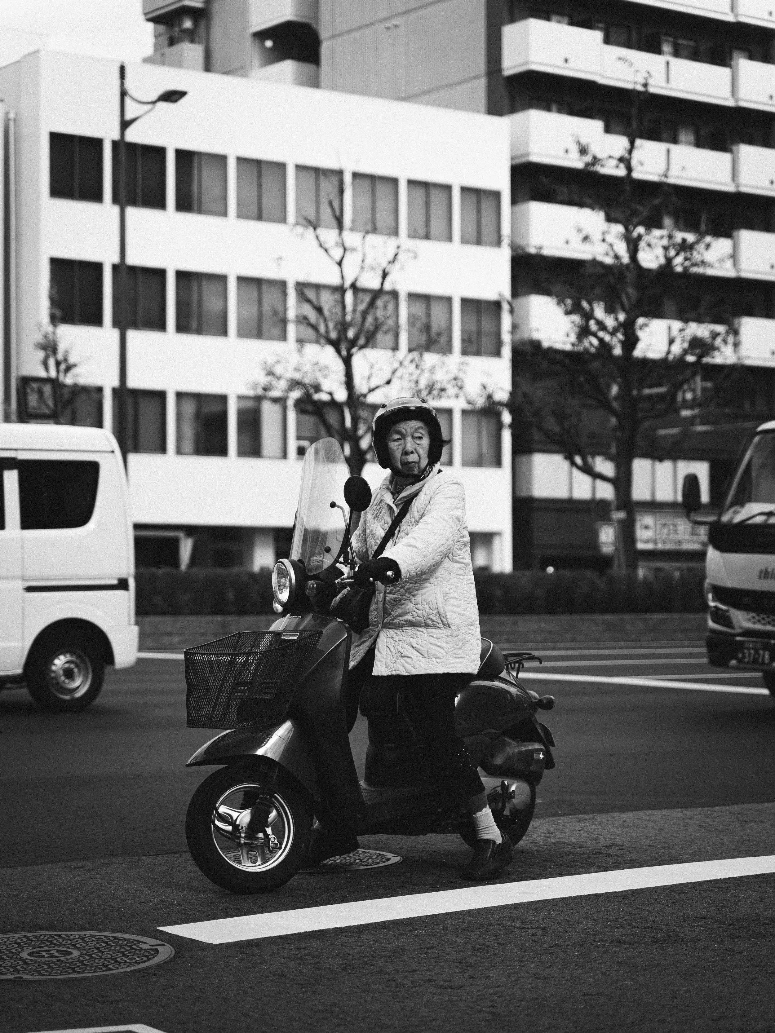An elderly woman wearing a helmet and quilted jacket, sitting on a scooter on a city street, with buildings, trees, cars, and streetlights in the background in black and white.