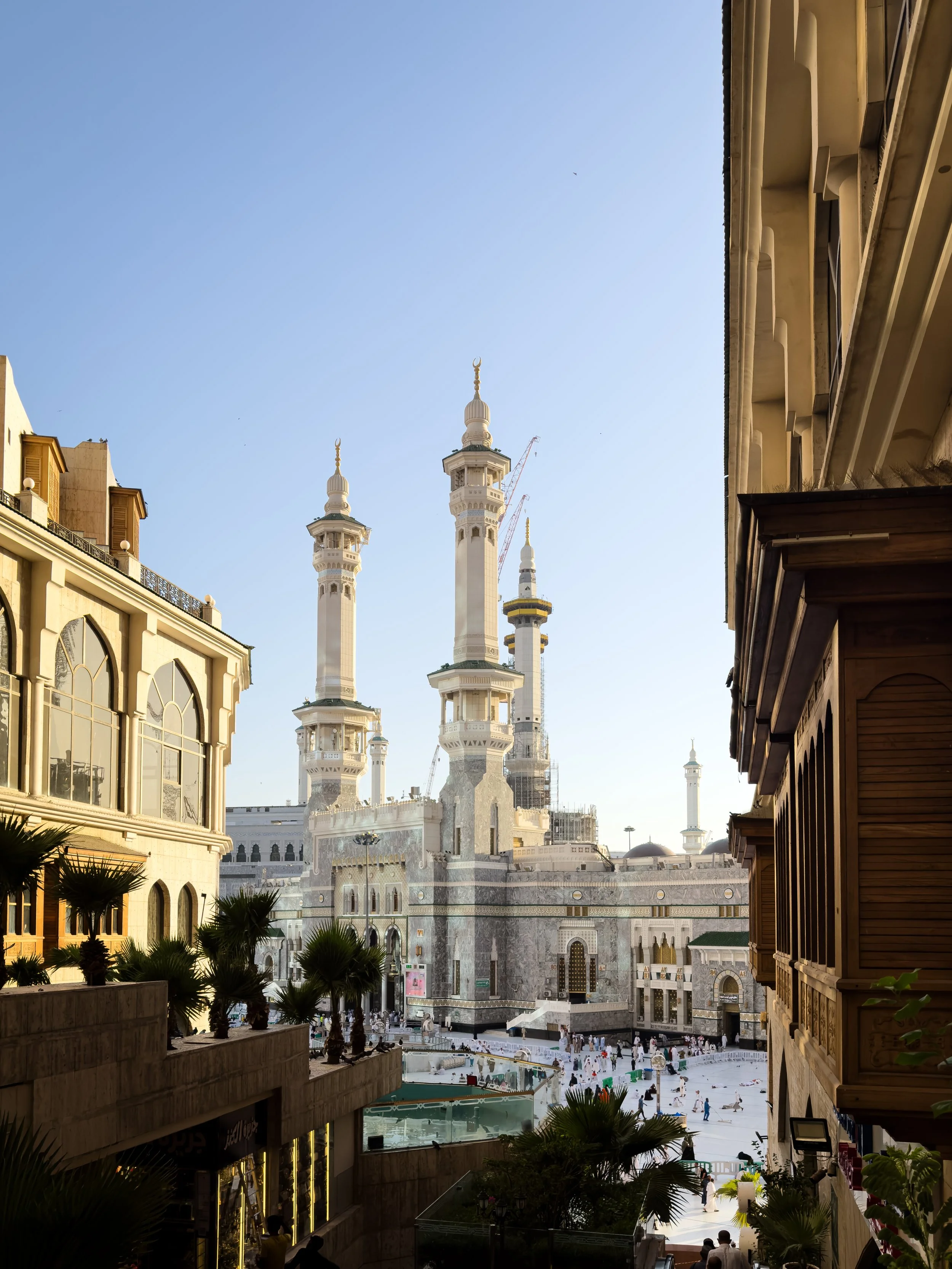 Masjidil Haram, Mecca, Saudi Arabia — The minarets rise into a clear blue sky at golden hour, pilgrims moving through the streets below. Even the approach feels like arrival.