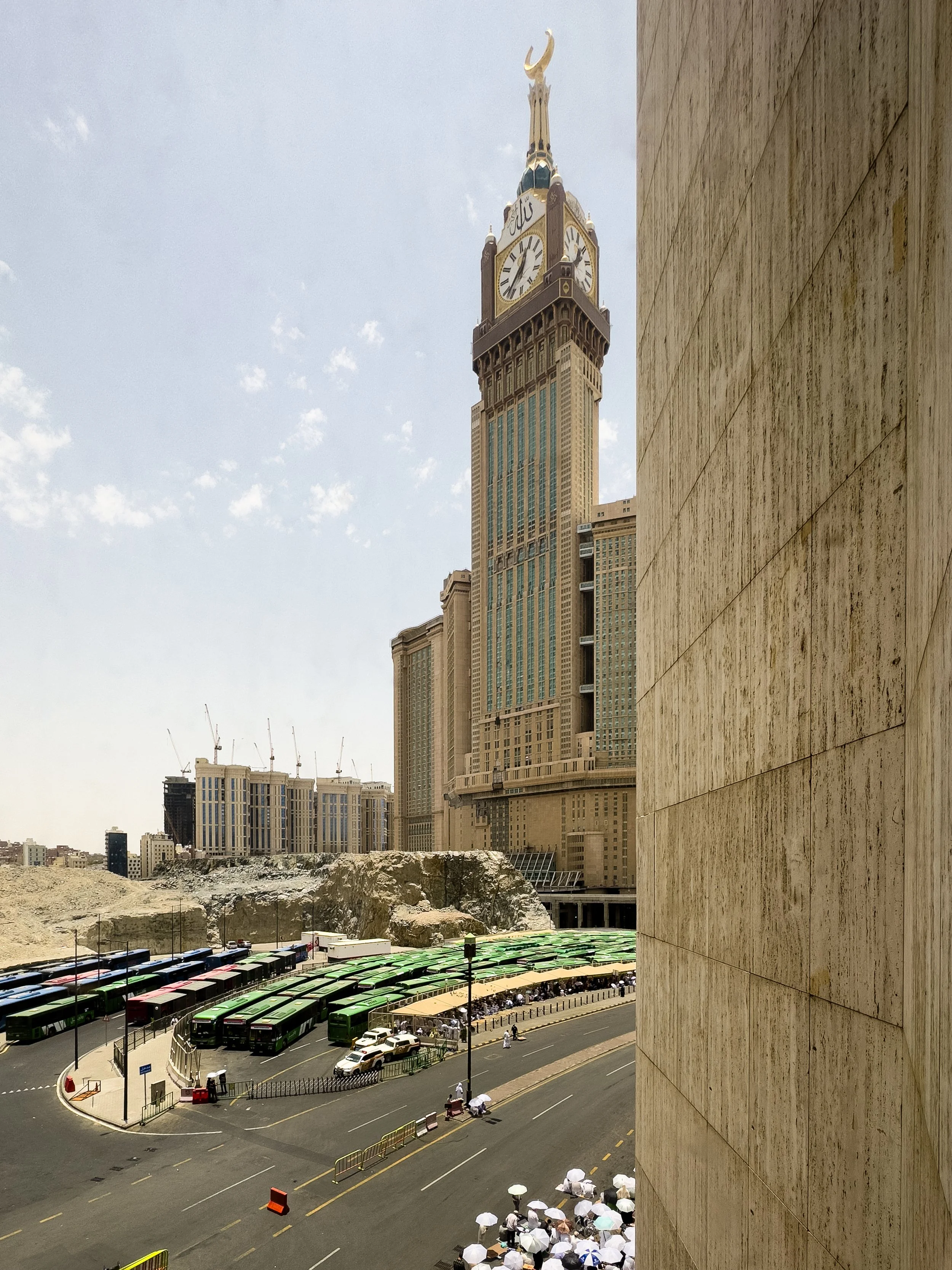 Makkah Royal Clock Tower, Mecca, Saudi Arabia — The Abraj Al-Bait clock tower rises above the Masjidil Haram, buses and pilgrims filling the streets below in the midday heat. The ancient and the monumental, side by side — Mecca as it is today.