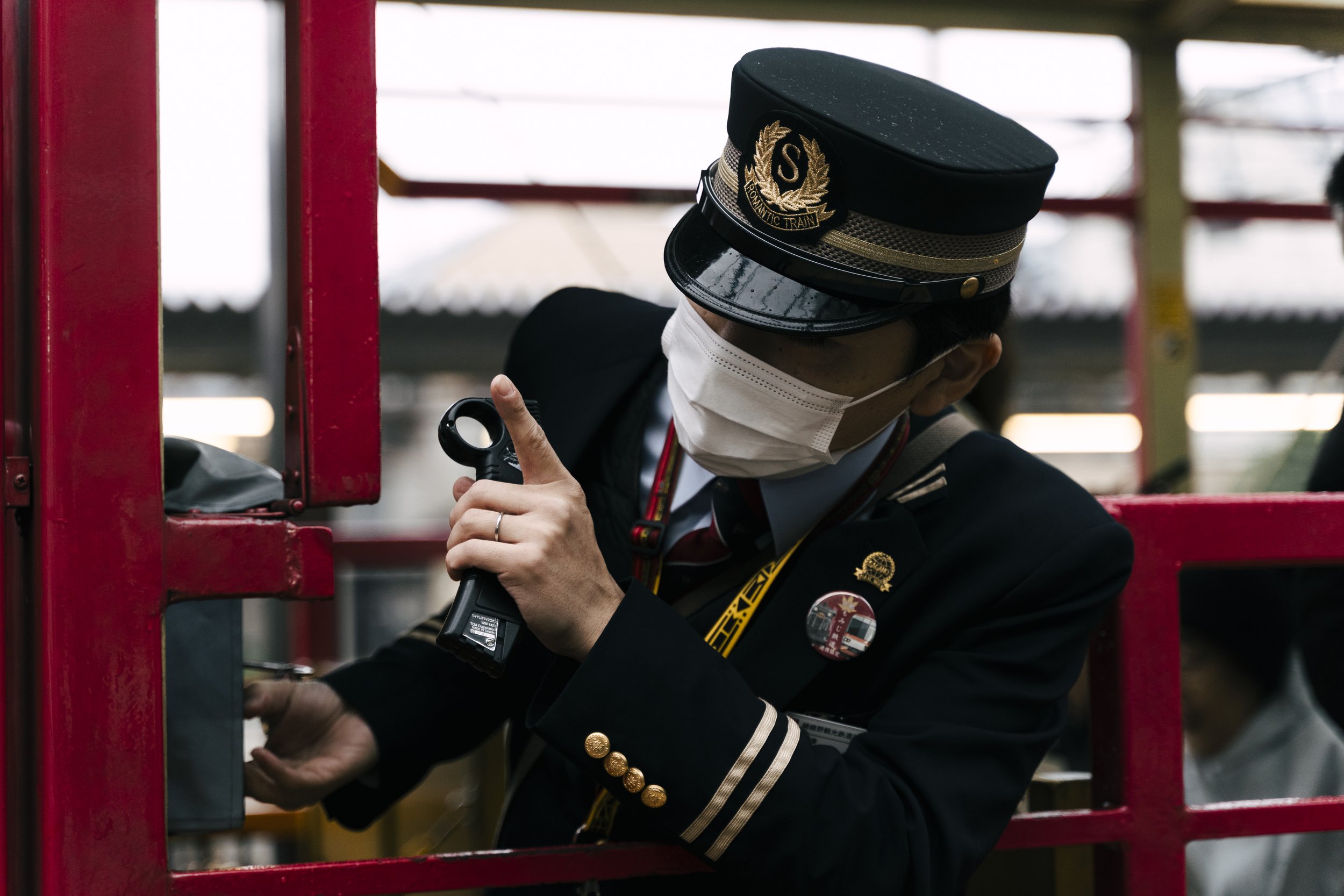 A person wearing a black hat with gold embroidery and a gold badge, a face mask, and a uniform with gold buttons, is adjusting or inspecting something near a red railing or structure, with a blurred background.