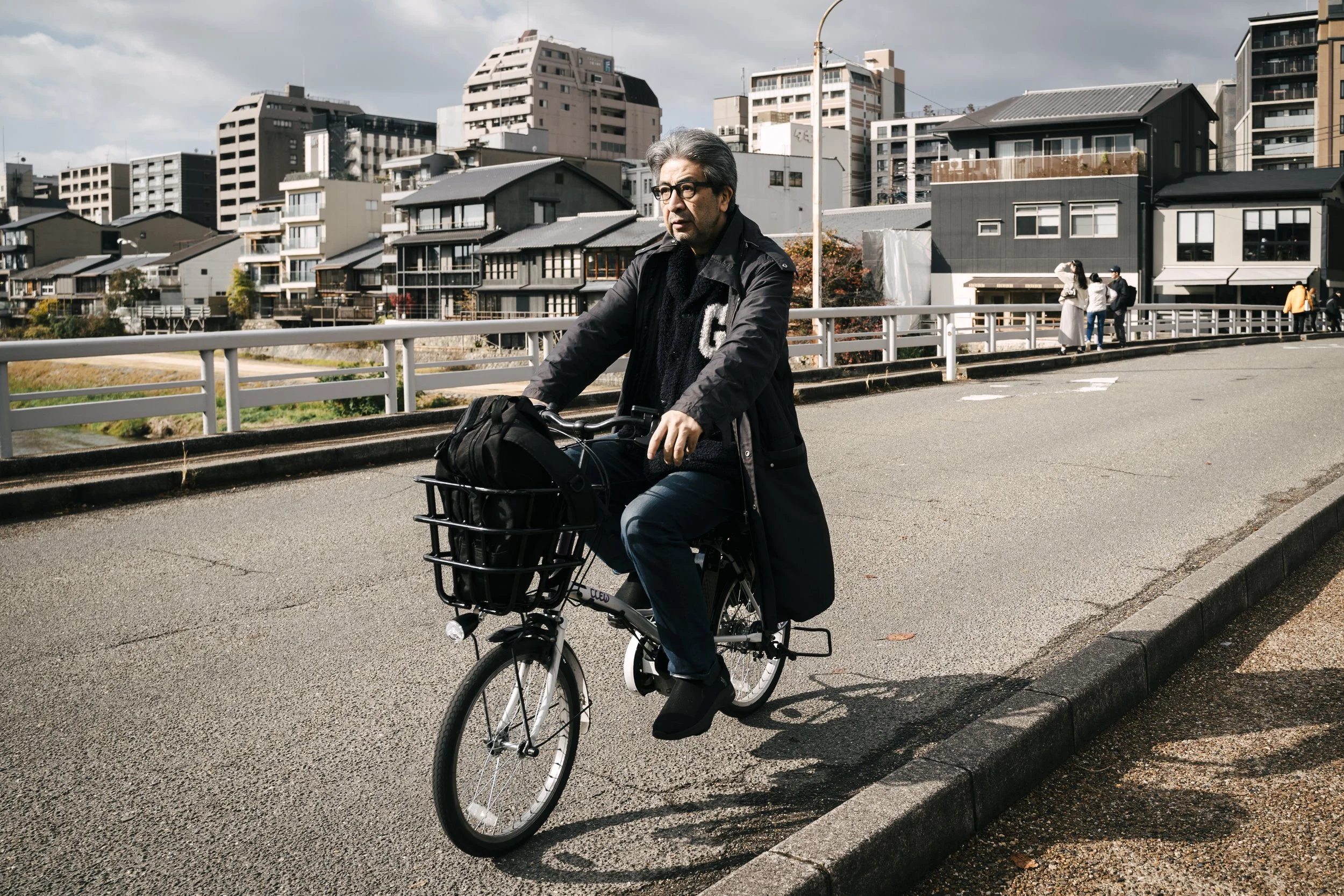 An older man with gray hair, glasses, and a serious expression riding a bicycle on a city street. He is dressed in a black jacket and dark pants, and there is a shopping bag in the bicycle's front basket. In the background, there are several modern a