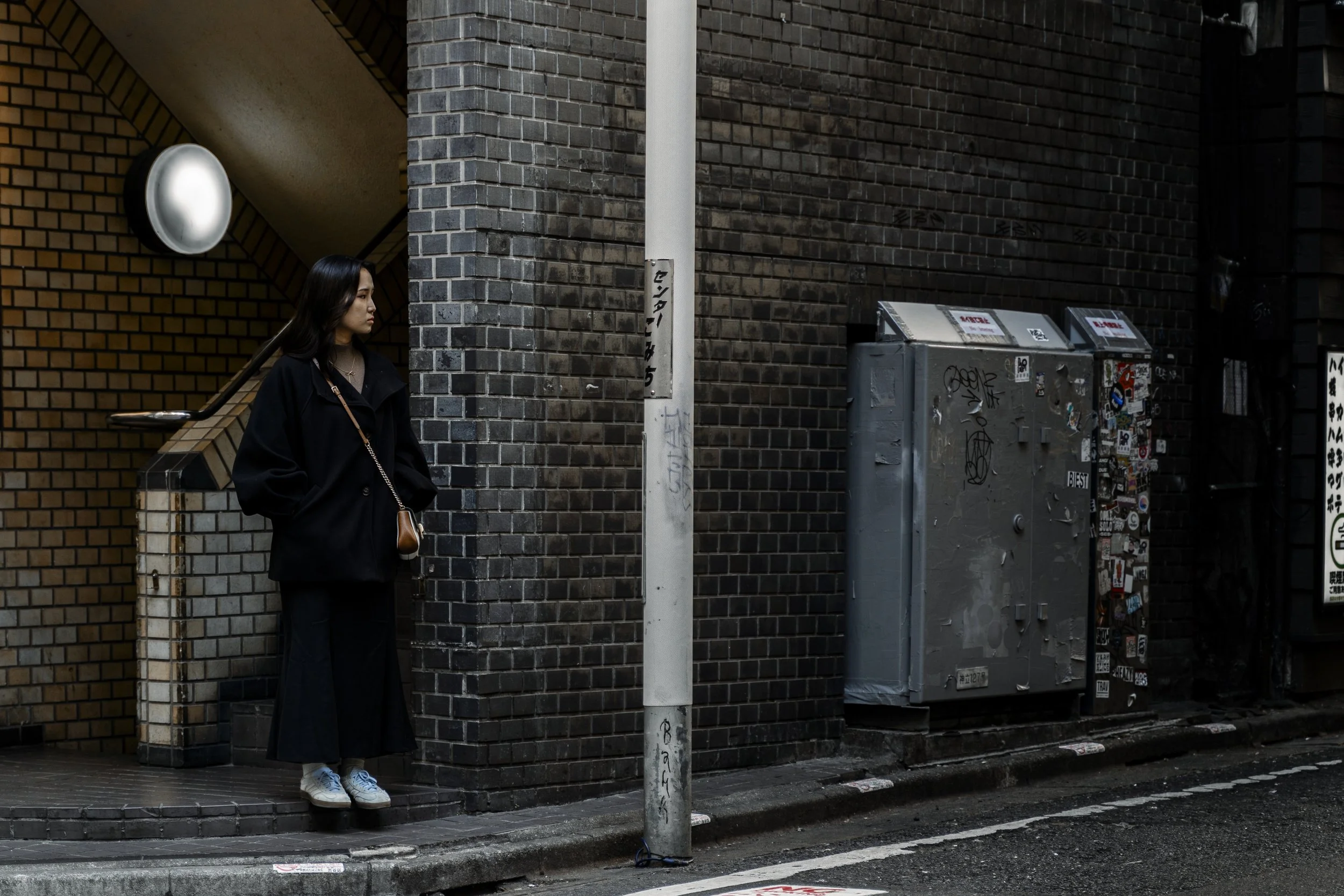 A woman with black hair, wearing a black coat, white sneakers, and carrying a brown bag, stands on a city sidewalk next to a brick wall, near utility boxes covered in street art and graffiti, with a metal pole in the foreground.