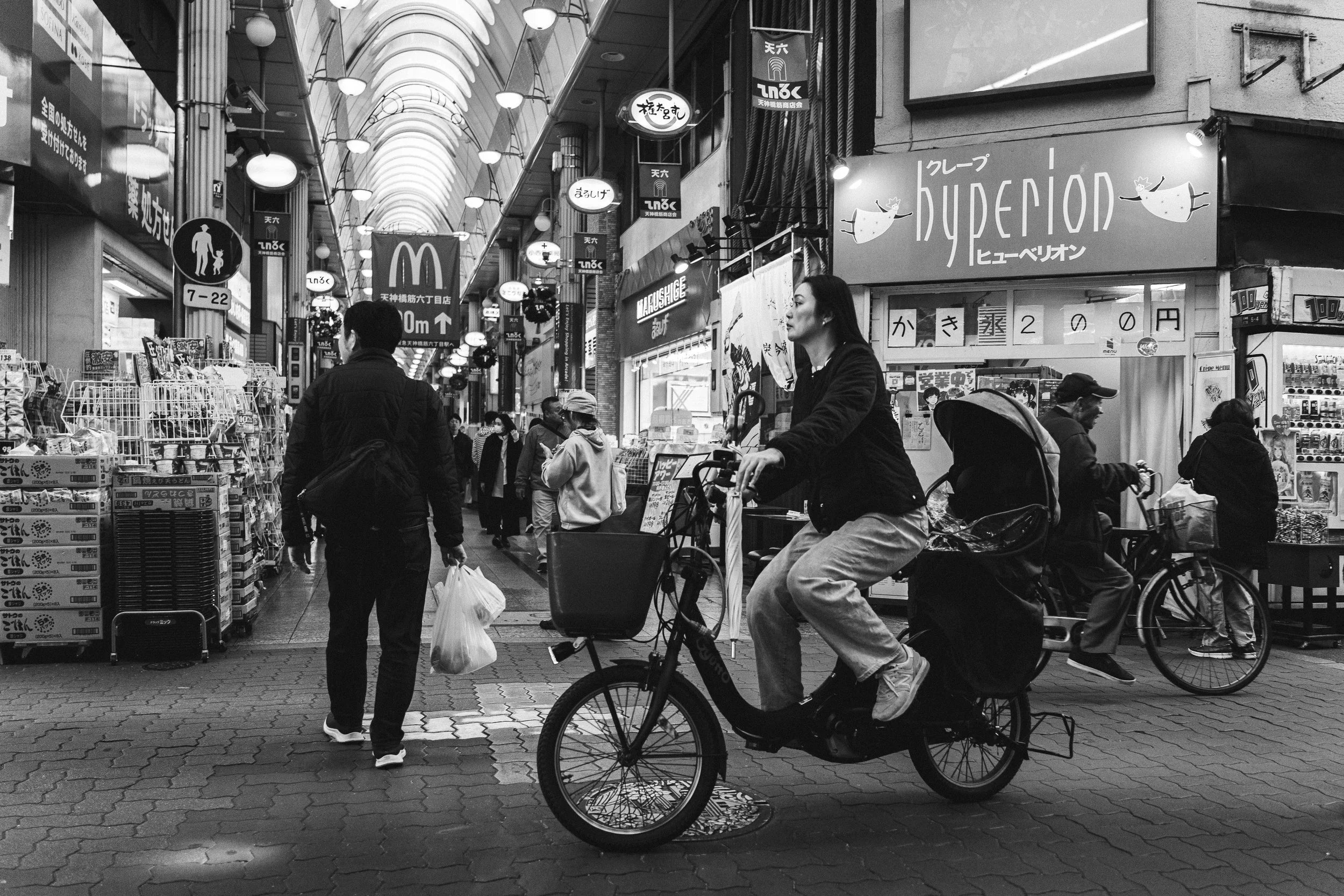 People shopping and riding bicycles inside a covered shopping arcade, with signs in Japanese, a McDonald's logo, and various stores.
