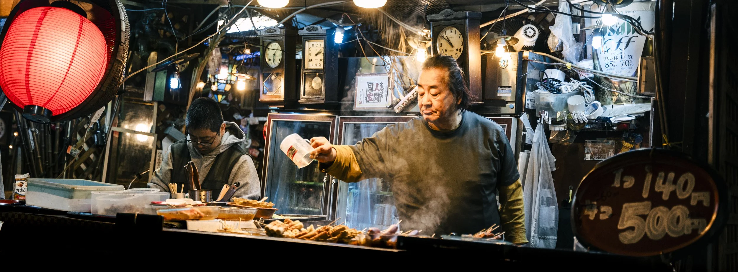 A street food vendor prepares skewered grilled food at a night market stall, with a woman sprinkling seasoning and a young man focused on cooking, surrounded by hanging lights, clocks, and various kitchen items, in a busy, cluttered environment.