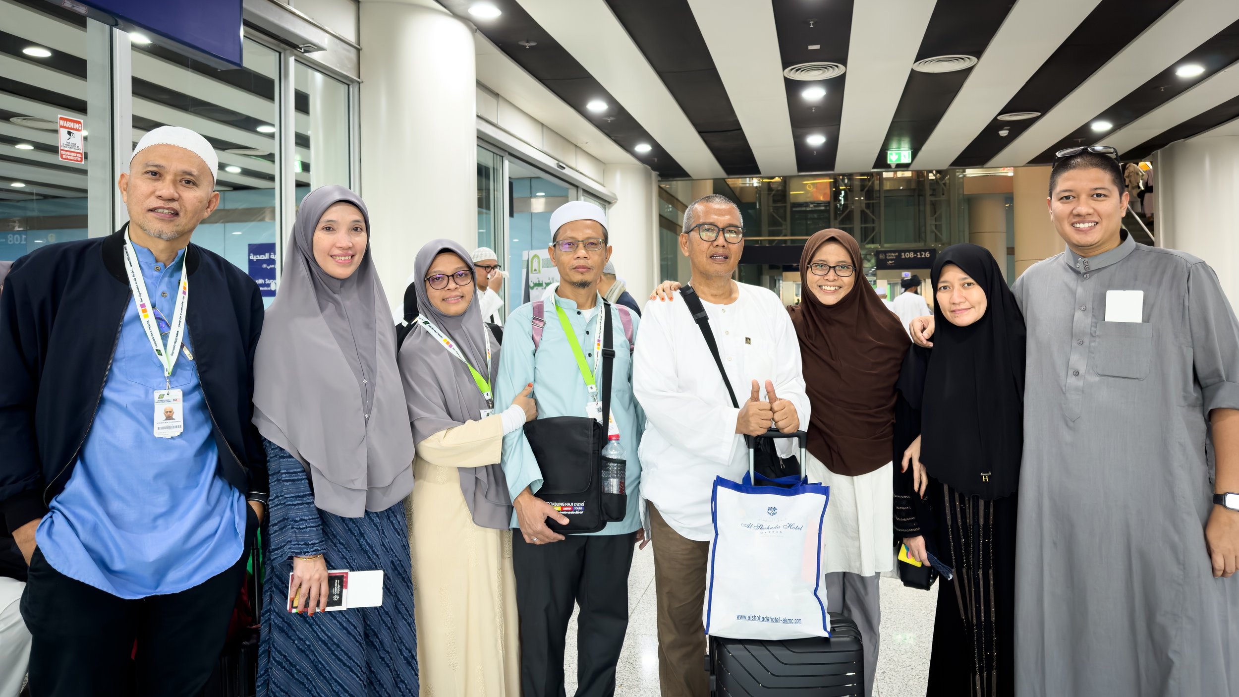 Group of nine diverse people standing together at an airport terminal, smiling and posing for the photo, with luggage and travel bags.