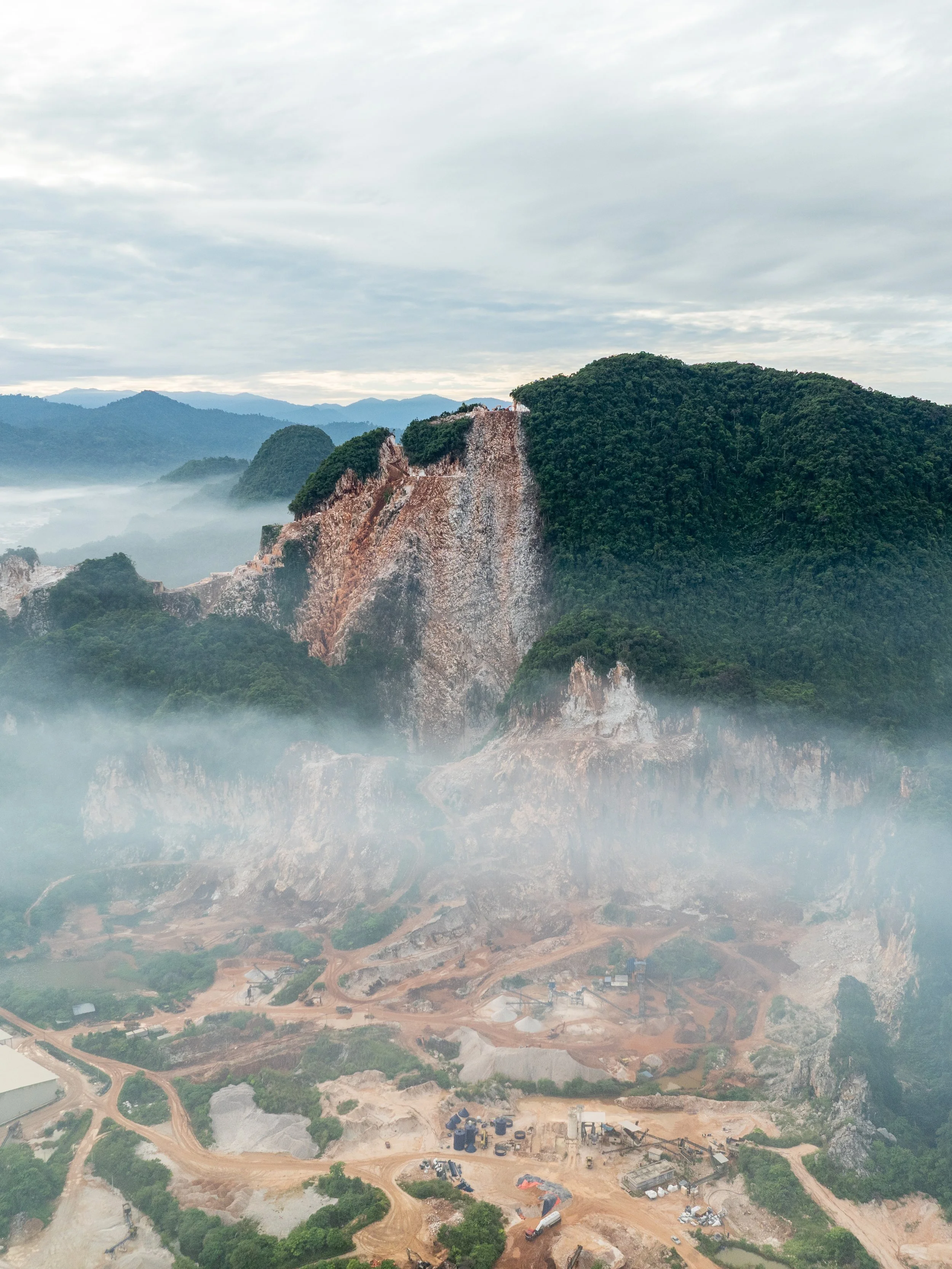 Morning mist veils an active quarry site at the base of a limestone hill in Ipoh, Perak, blurring the boundary between industry and nature.