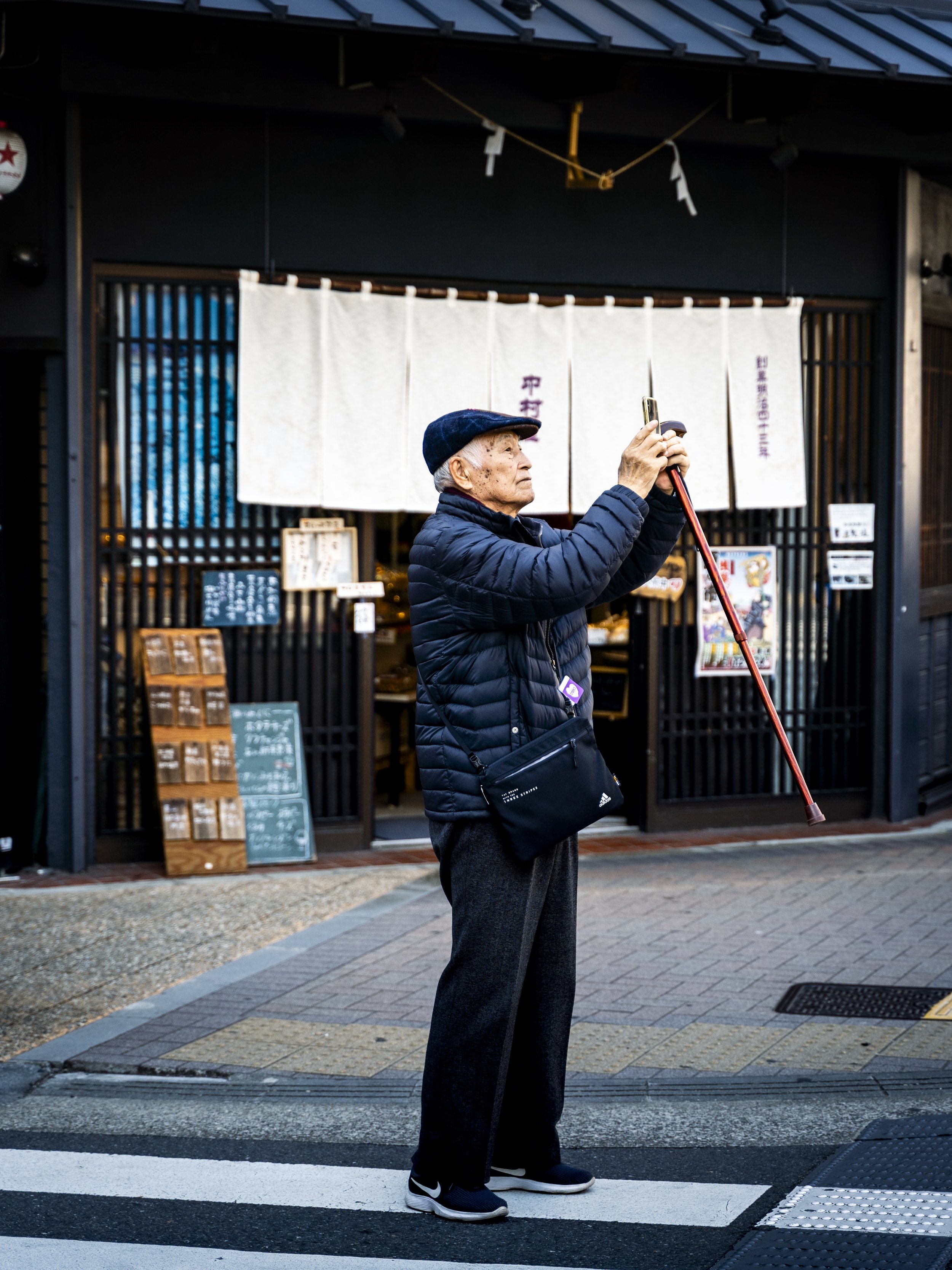 An elderly man in a blue puffer jacket and black pants stands at a crosswalk, holding a red cane upright, with a store featuring Japanese signage and a traditional hanging curtain in the background.