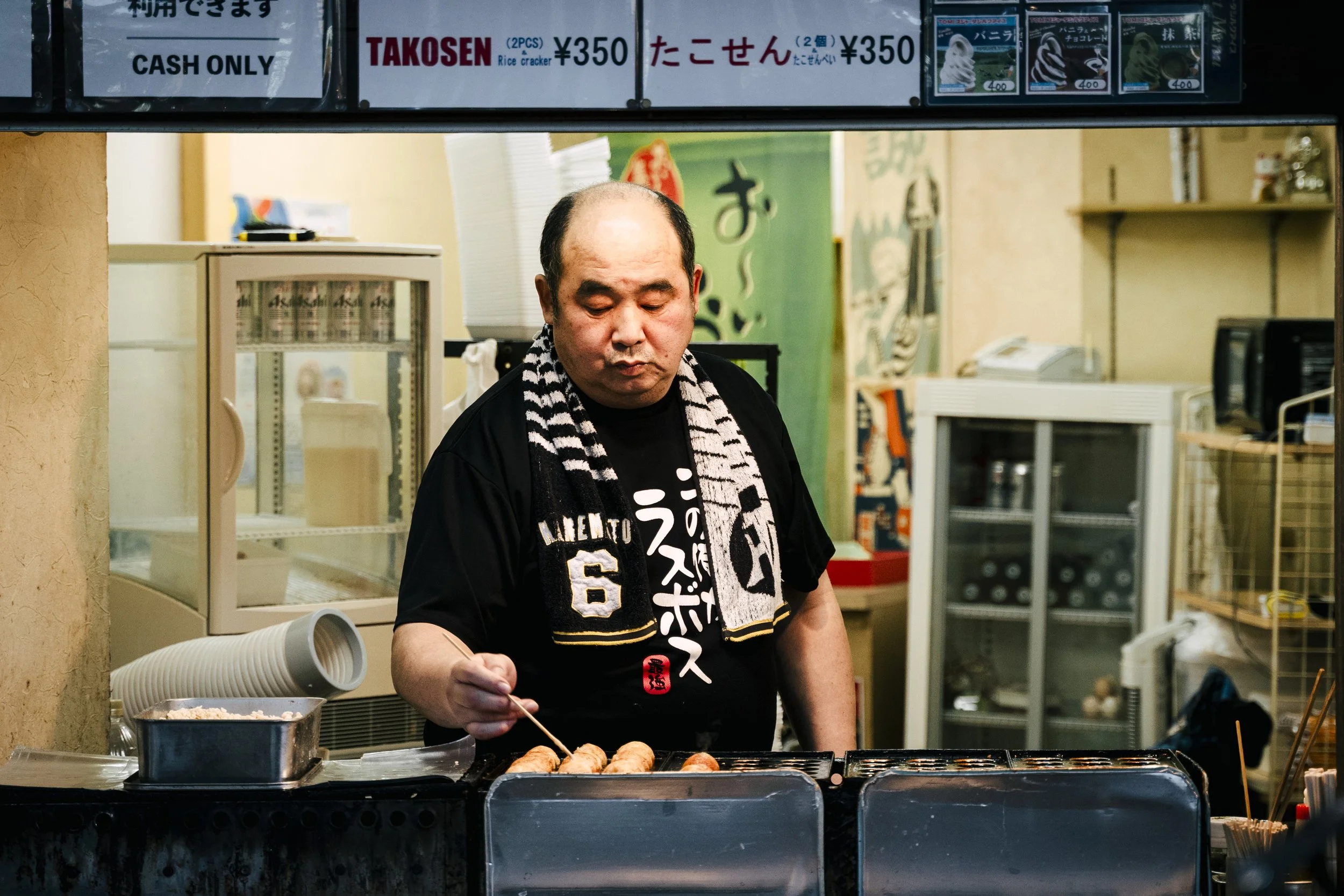 A man cooking takoyaki, a Japanese snack, at a food stall. He is using a skewer to turn the round batter balls on a griddle. The background shows various signs with Japanese writing and some packaged ice cream or dessert products.