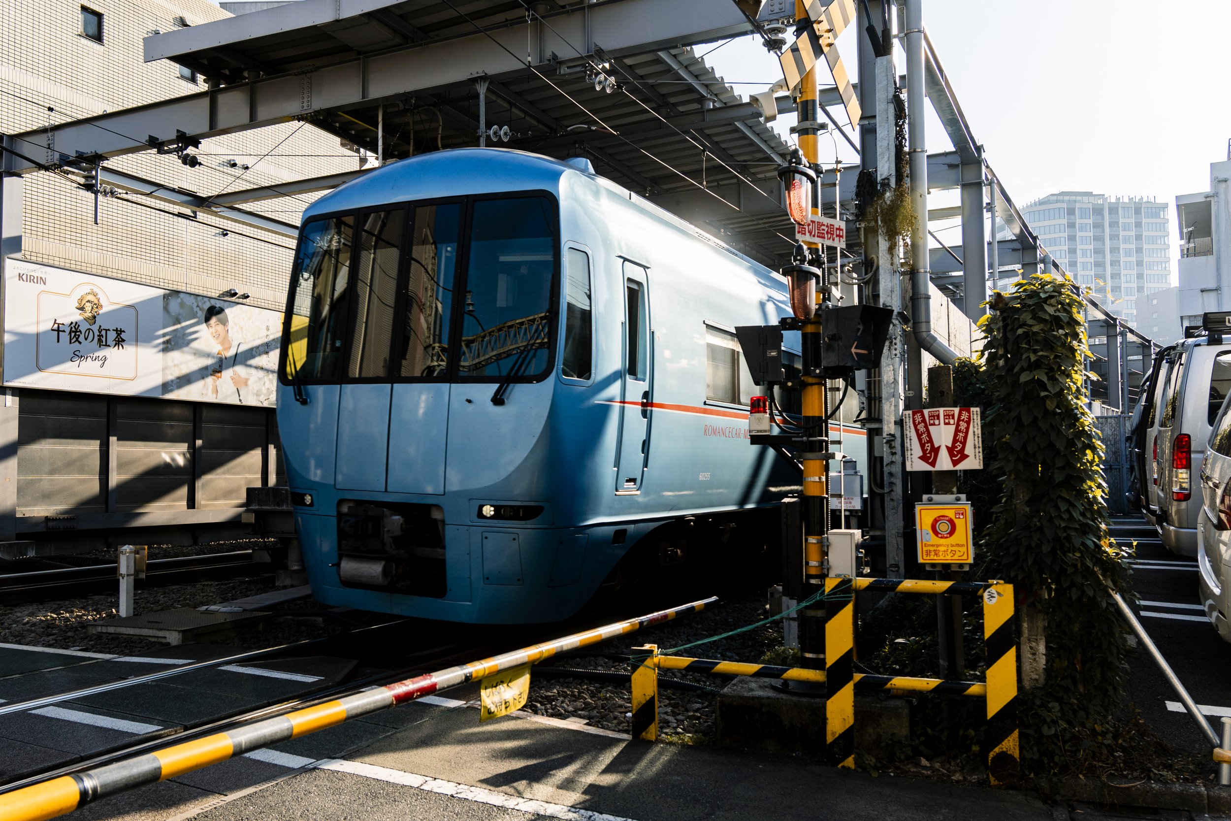 Odakyu Romancecar GSE, Tokyo — The vivid blue GSE clears a level crossing, barriers down, cars queued on both sides. A luxury limited express squeezing through the ordinary machinery of a Tokyo street.