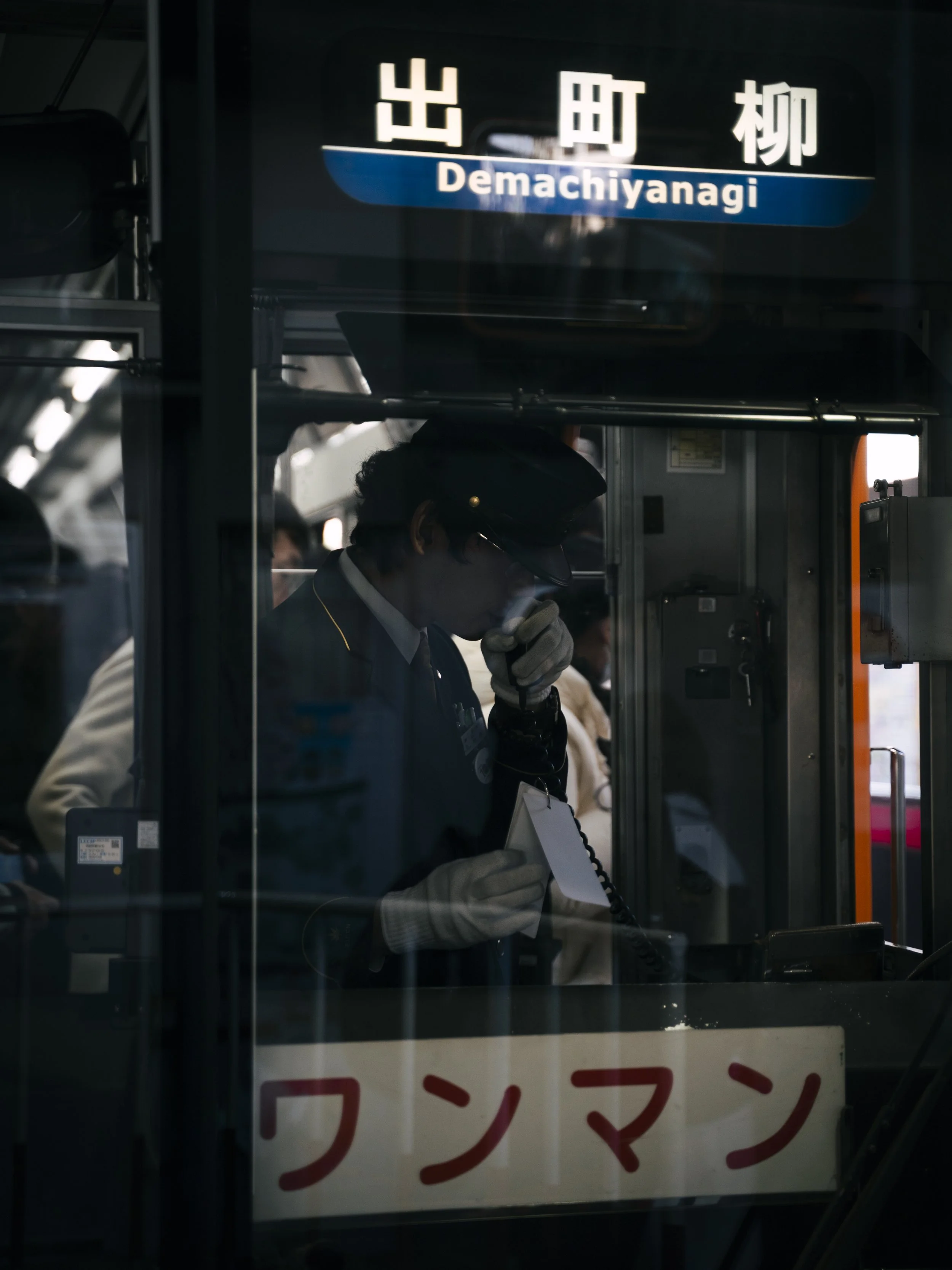 A train station sign displaying the name 'Demachiyanagi' in both Japanese characters and English. A person, dressed in a uniform and headphones, is speaking into a microphone inside a ticket booth or station office, partially visible through the wind