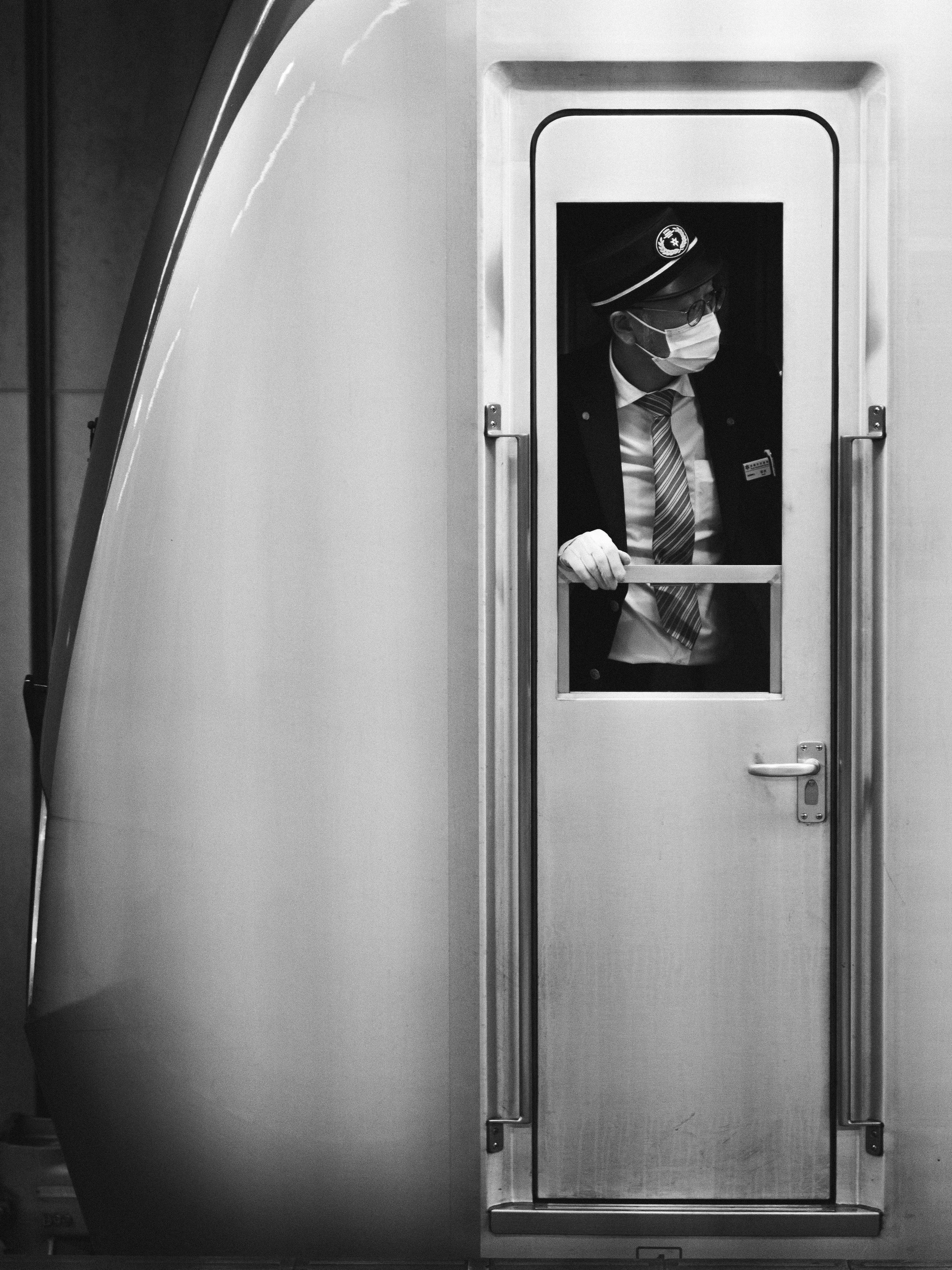 A man wearing a hat, glasses, and a face mask is standing inside a small, enclosed space with a window, looking out.