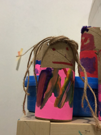 Handmade craft figure made from a cardboard tube, decorated with paint, yarn hair, and paper features, placed on a wooden surface.