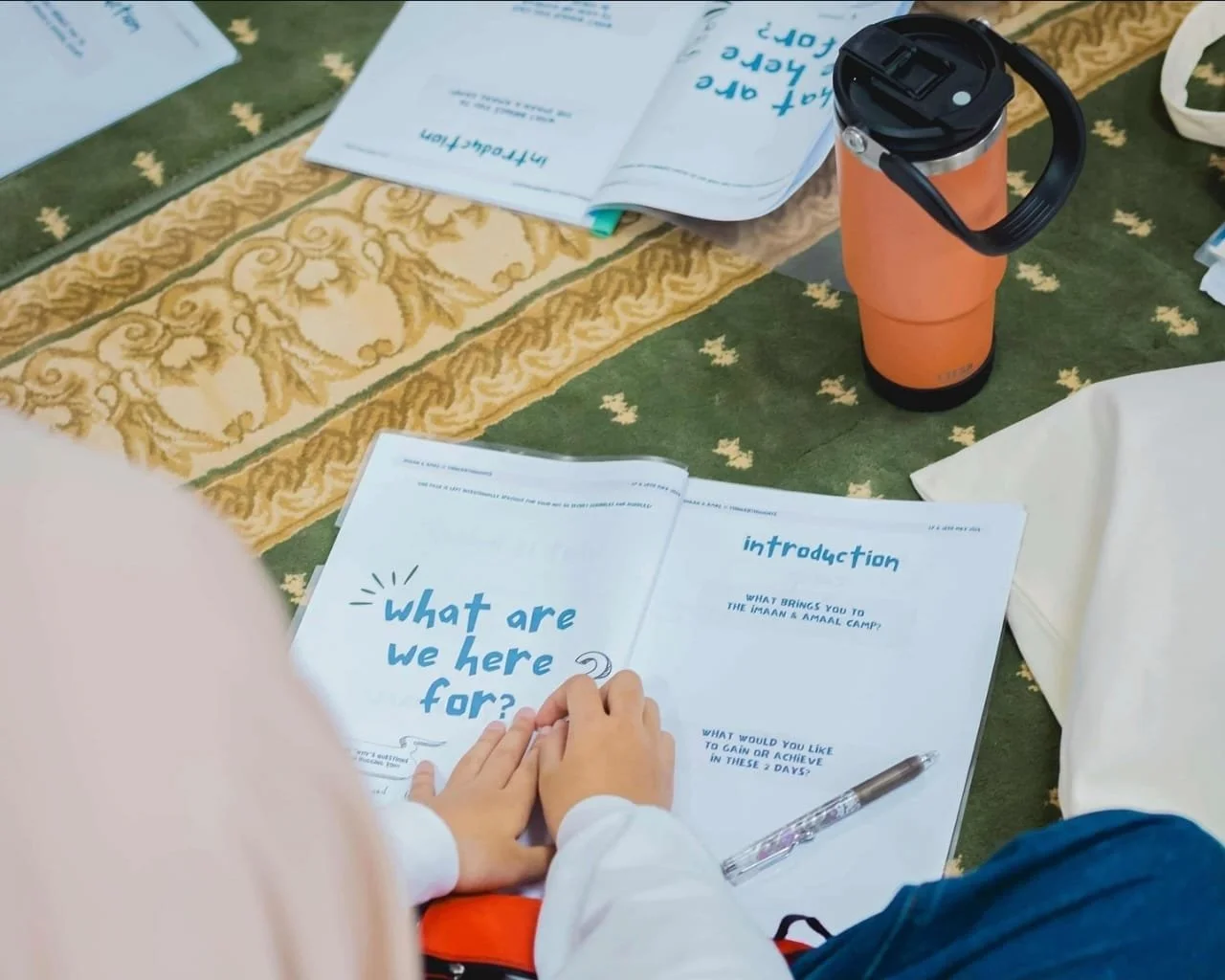 Open booklet on a green and gold floral tablecloth with the question, "what are we here for?" a hand holding a pen is visible, along with a water bottle, papers, and a white bag.