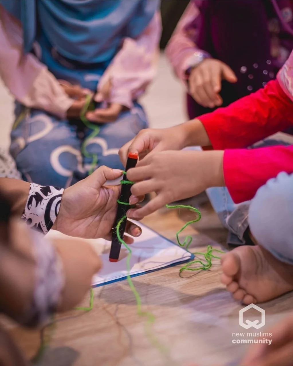People gathered around a table, working together on a project involving string, with one person holding a marker and green string, in a collaborative activity.