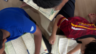 Two children reading Quranon the floor, one in a blue shirt and the other in a red maroon shirt.
