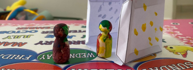 Two small figure toys with painted colors, one predominantly purple and the other yellow and green, positioned on a colorful board game surface. There is a paper bag with yellow markings and some background objects.