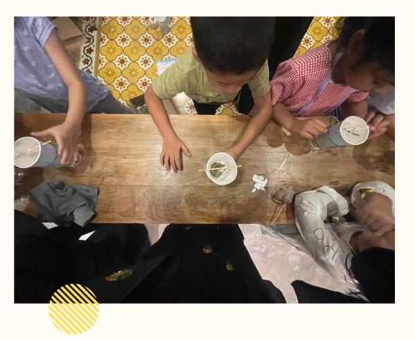 Children sitting around a wooden table, each holding a cup with some liquid and straws inside, in a room with yellow patterned floor tiles.