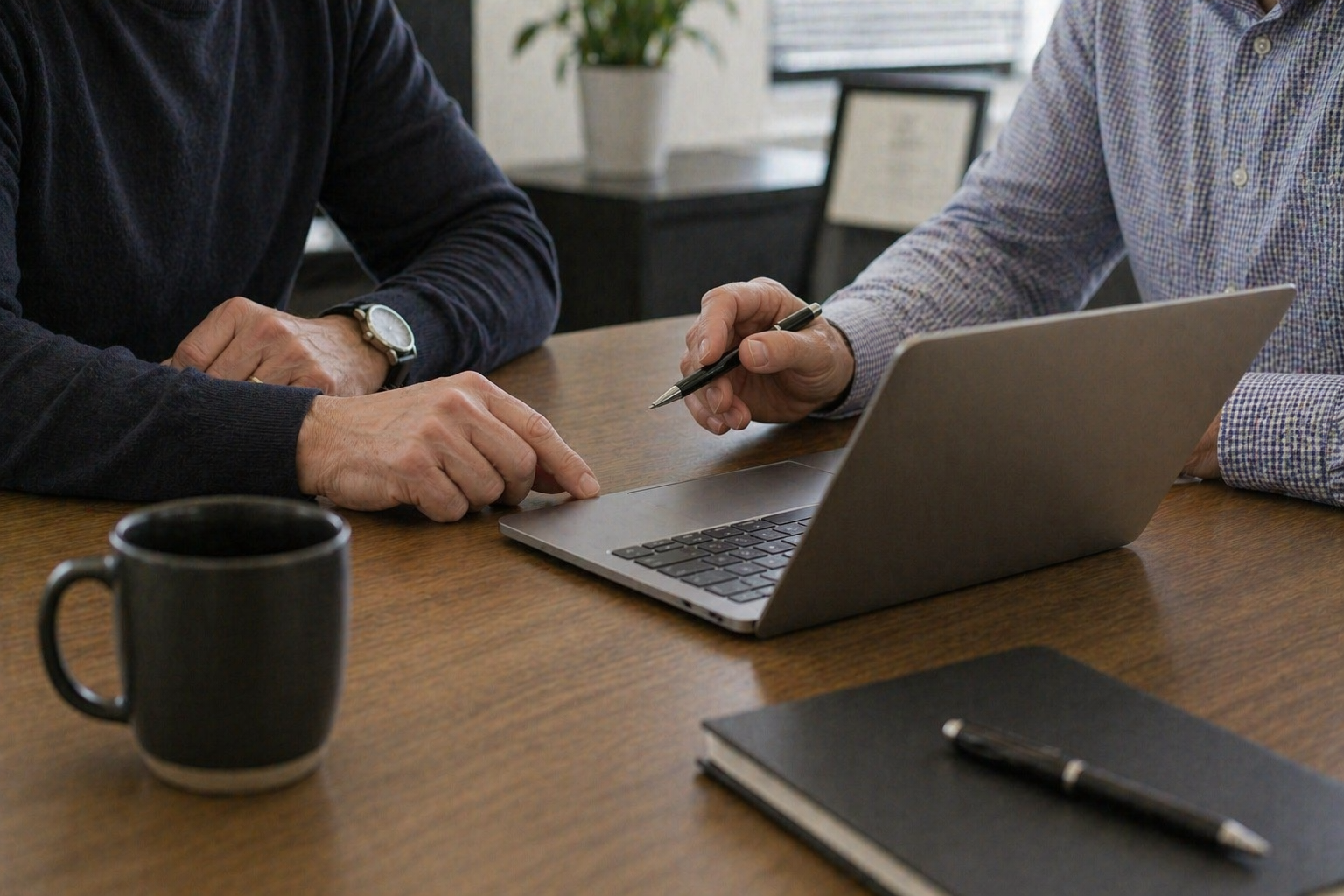 Two people in a business meeting pointing at a laptop screen on a wooden table, with a coffee mug, a notebook and pen, and a potted plant in the background.