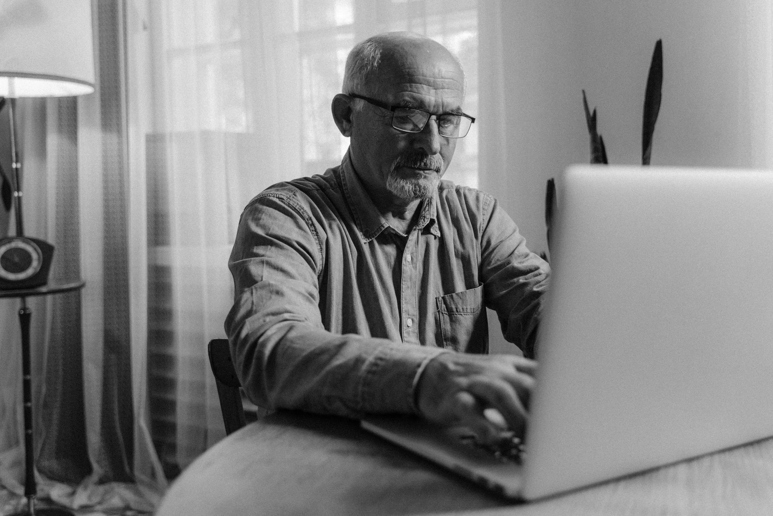 An older man with glasses and a beard, sitting at a desk, working on a laptop in a well-lit room with large windows, sheer curtains, a lamp, and a vintage telephone on a side table.