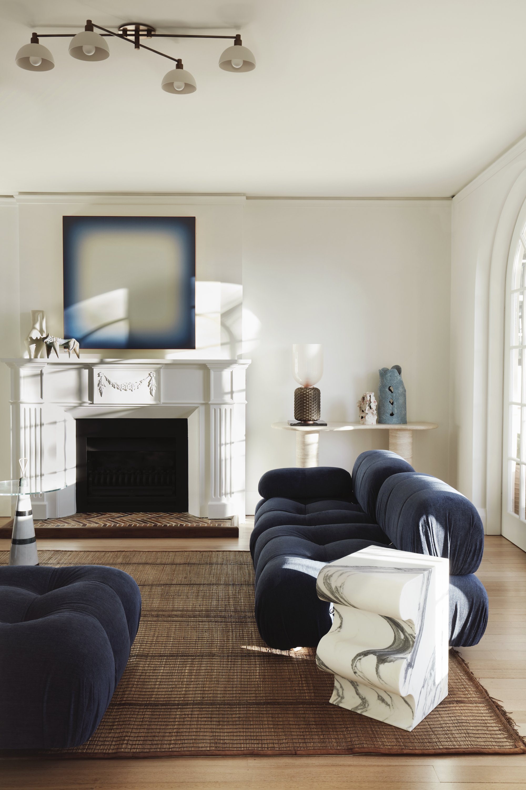 A living room with a white fireplace and mantel, decorated with small animal figurines. There is a large abstract painting above the fireplace, a marble and black side table with a lamp, and several plush dark blue chairs arranged around a brown area rug. Natural light fills the room from a large window to the right.