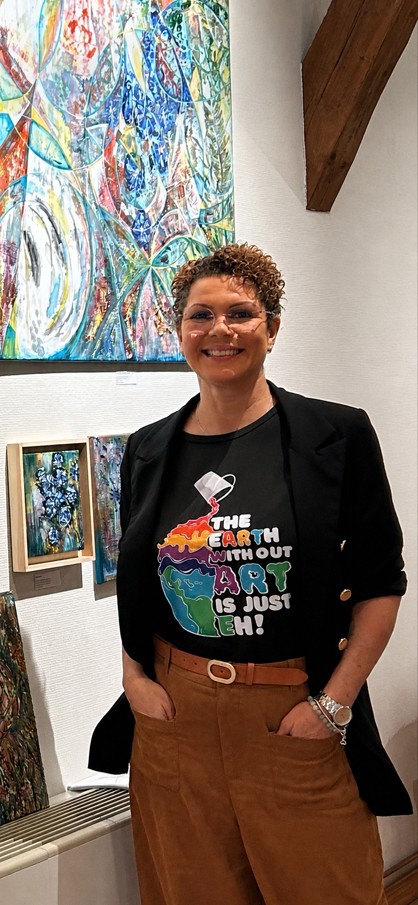 A woman with short curly hair, glasses, and a bright smile standing in an art gallery. She is wearing a black T-shirt with a colorful Earth and paint design and the words "The Earth Without Art Is Just Eh!", a black blazer, and brown high-waisted pants with a belt. Behind her, there are vibrant, abstract paintings displayed on the wall.